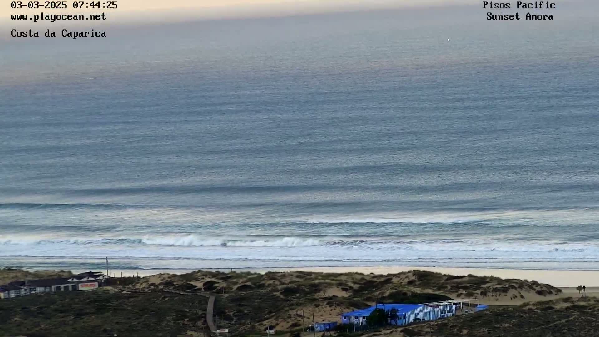 An ocean beach with rolling waves under a mostly clear, pale sky shows a sandy shore with some buildings and vegetation visible on the dunes.
