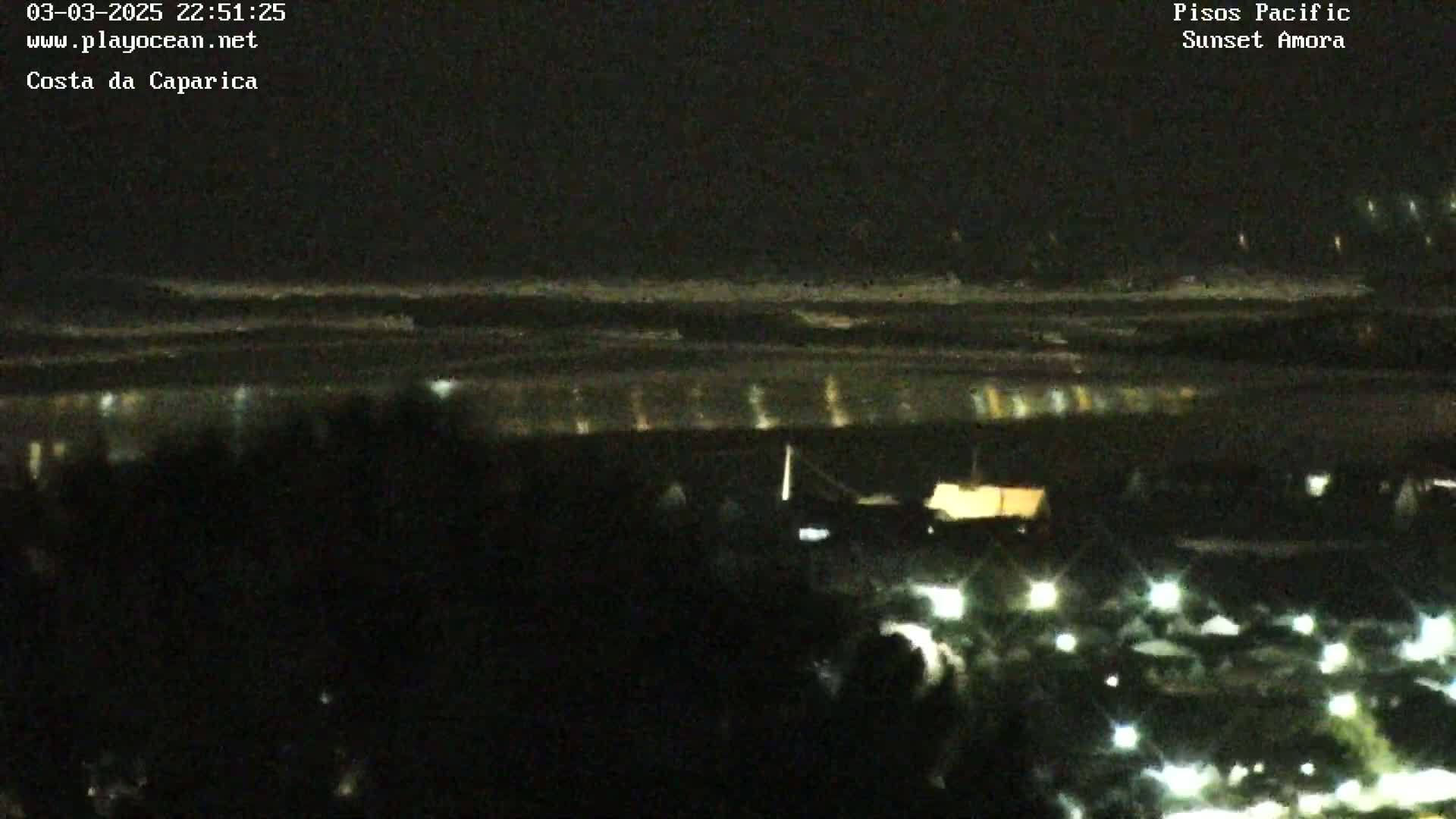 A nighttime, low-light view of a dark beach with a faint reflection of lights on the water and many small lights visible closer to the camera.