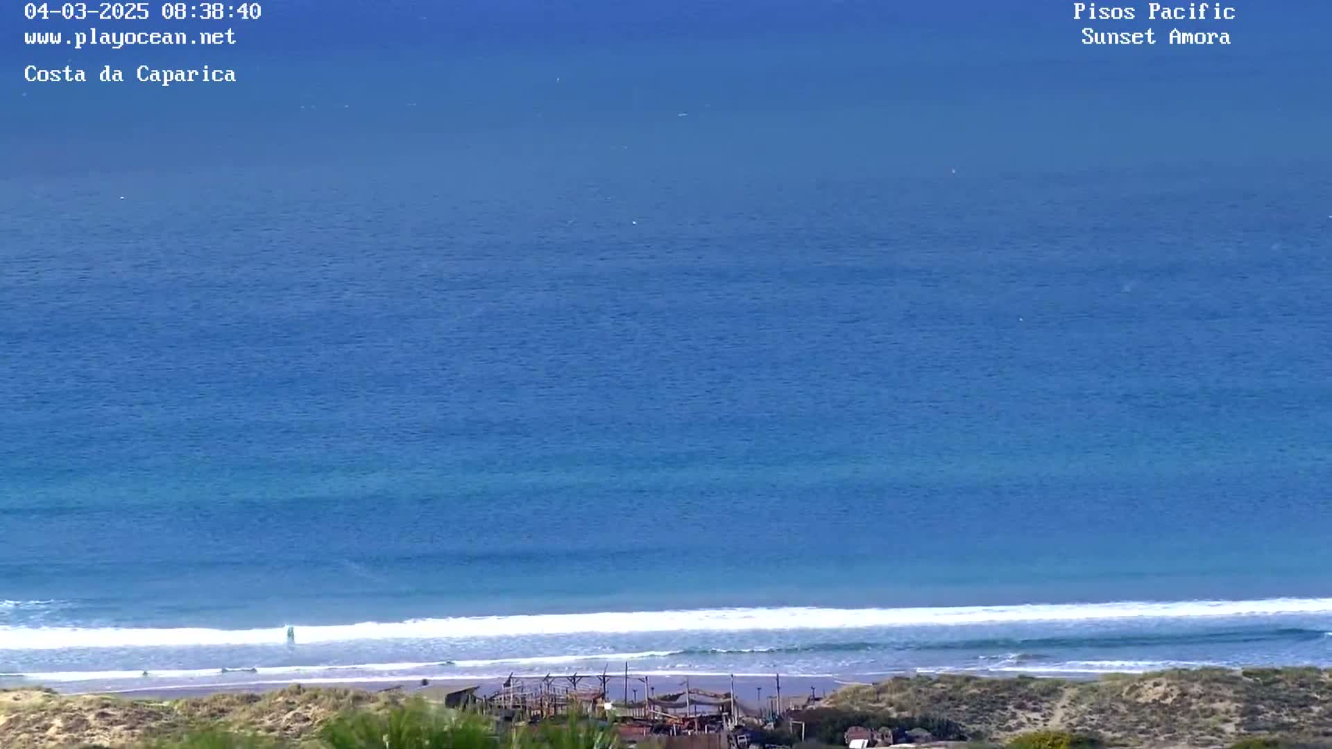 A high-angle view shows a calm ocean meeting a sandy beach with small structures visible near the shoreline on a clear day.