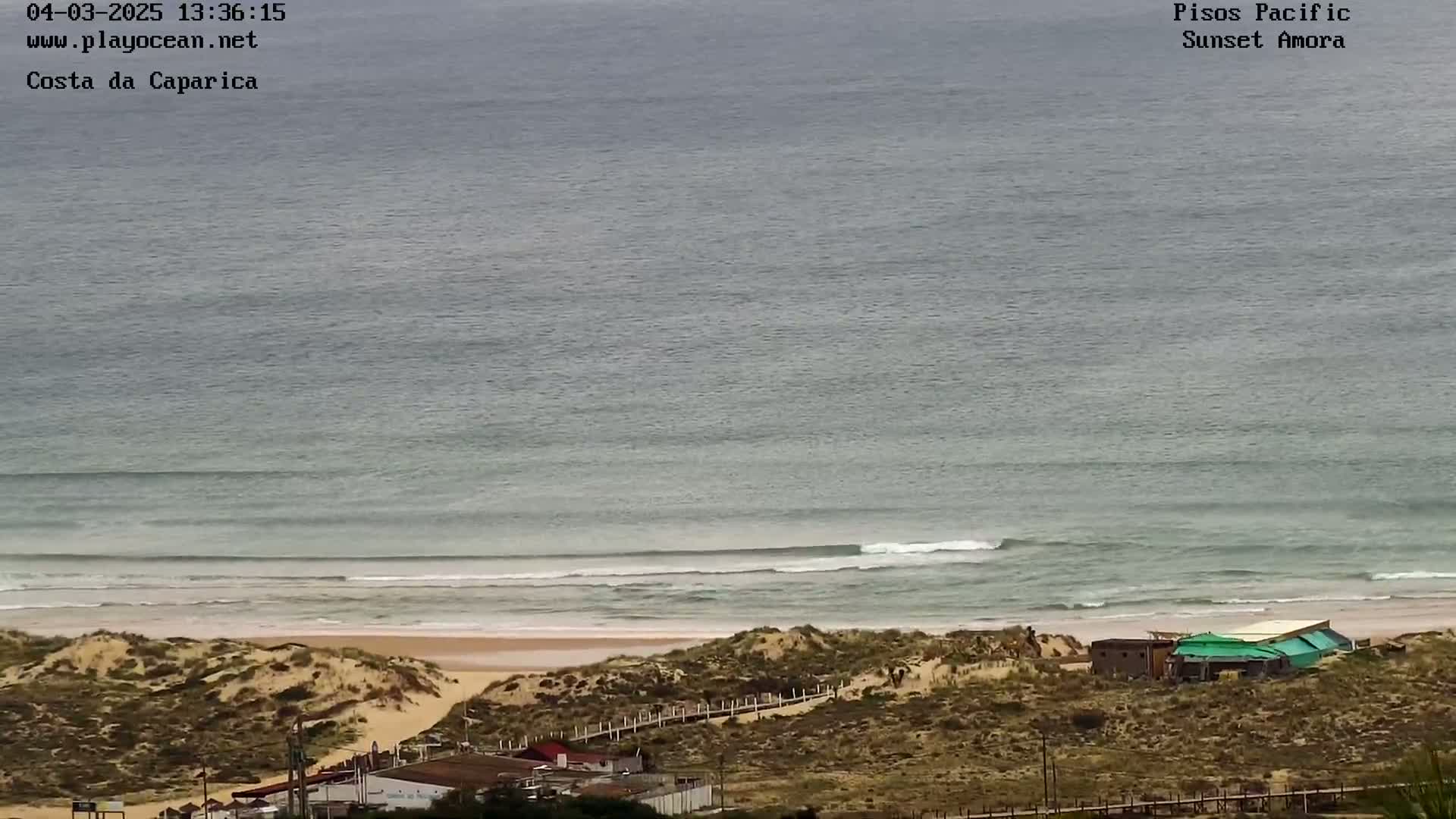A high-angle view shows a sandy beach with gentle waves rolling onto the shore under an overcast sky, with a few buildings visible near the dunes.