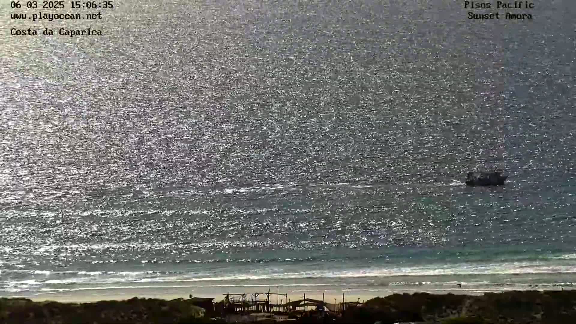 A distant boat is visible on a sparkling ocean under a bright sky, with a sandy beach and shoreline structures in the foreground.