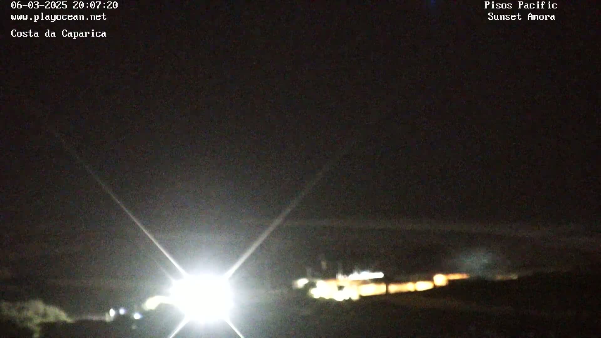 A nighttime long shot shows a bright light shining from the lower left corner of the frame, illuminating a dark landscape with faintly visible lights in the distance under a clear, dark sky.