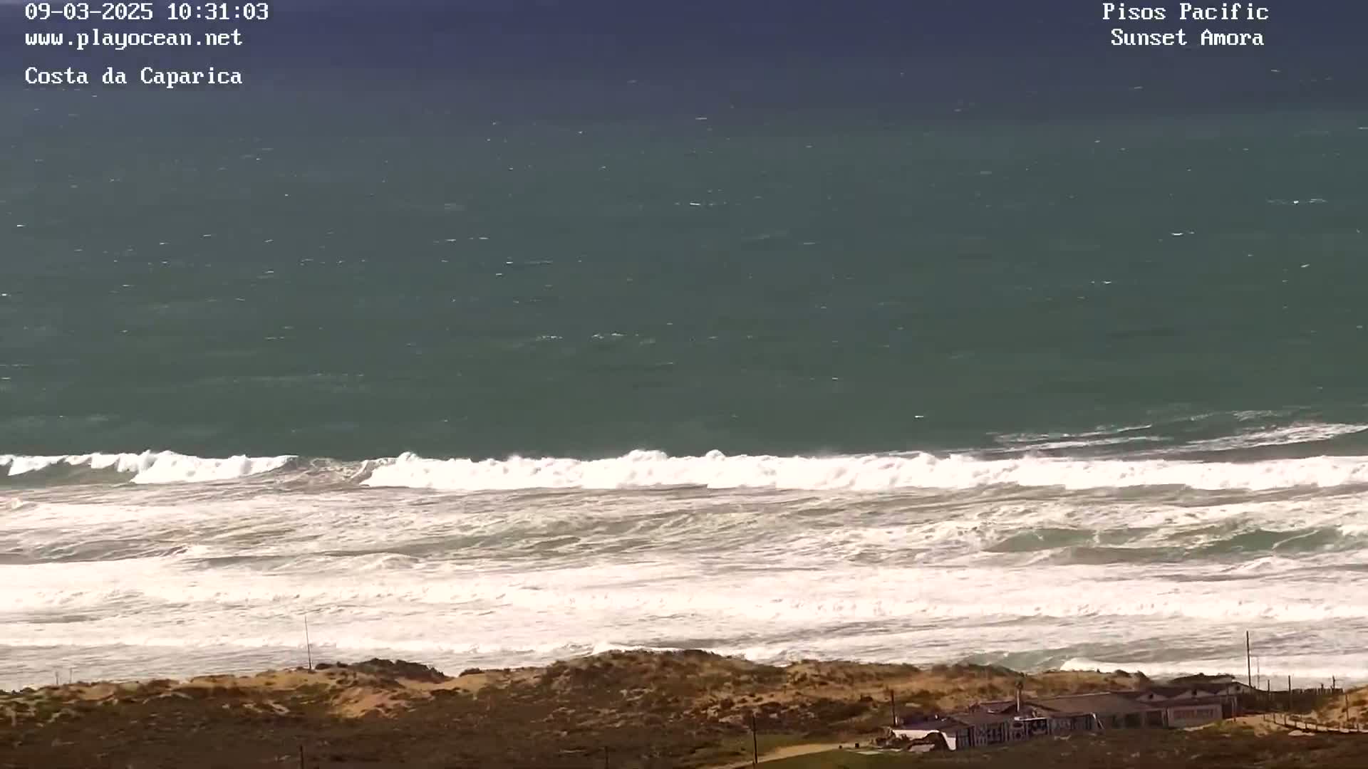 A high-angle view shows a stormy ocean with white-capped waves breaking on a sandy shore near some buildings.