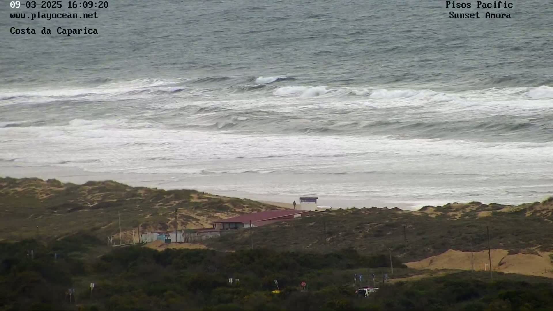 A sandy beach with a building and some vegetation is shown, overlooking a choppy ocean with waves breaking on the shore.
