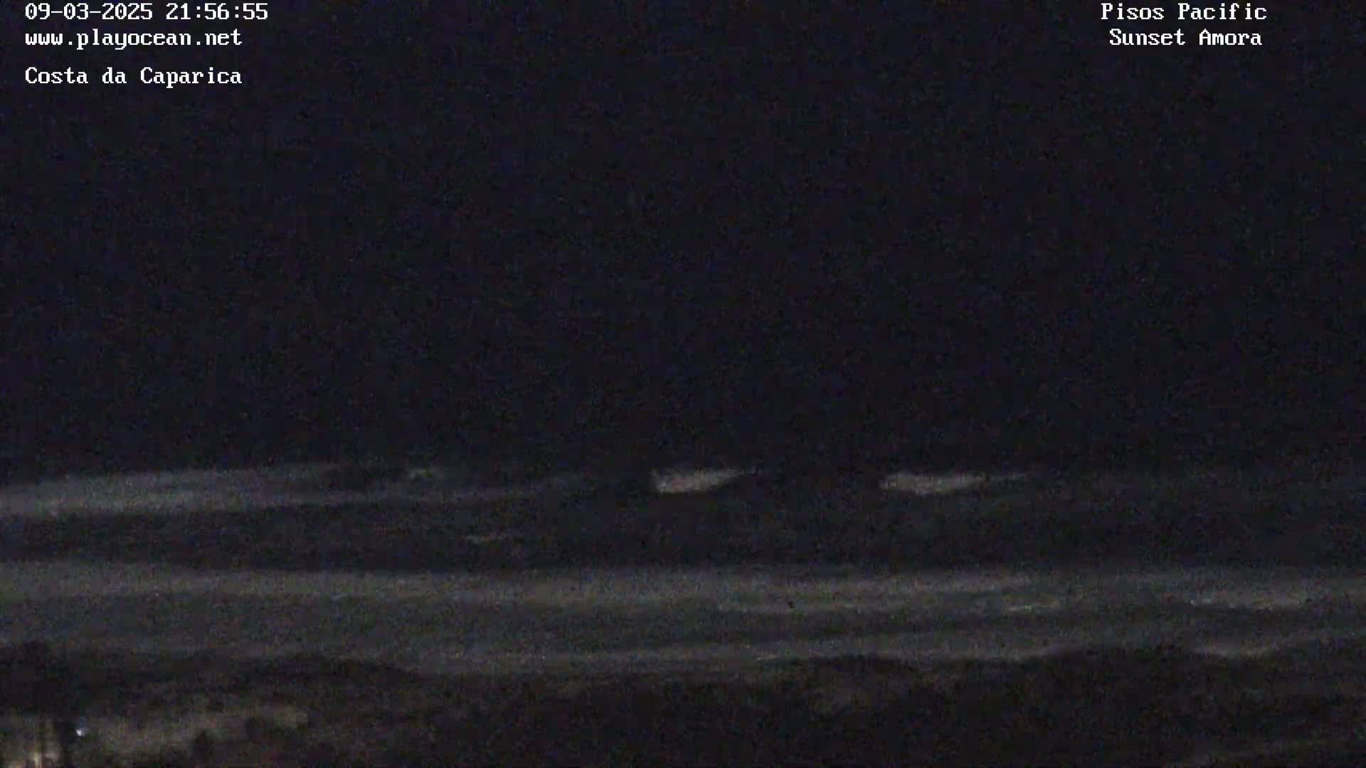 A dark, nighttime ocean scene shows faint waves breaking on a shoreline under a mostly clear sky.