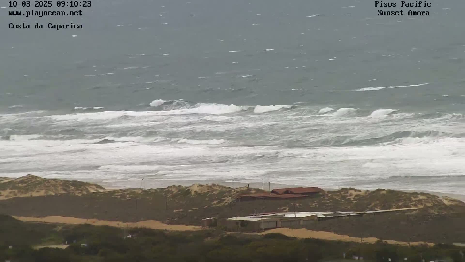 A high-angle view shows a beach with several buildings near the shore, and a turbulent ocean with white-capped waves under an overcast sky.