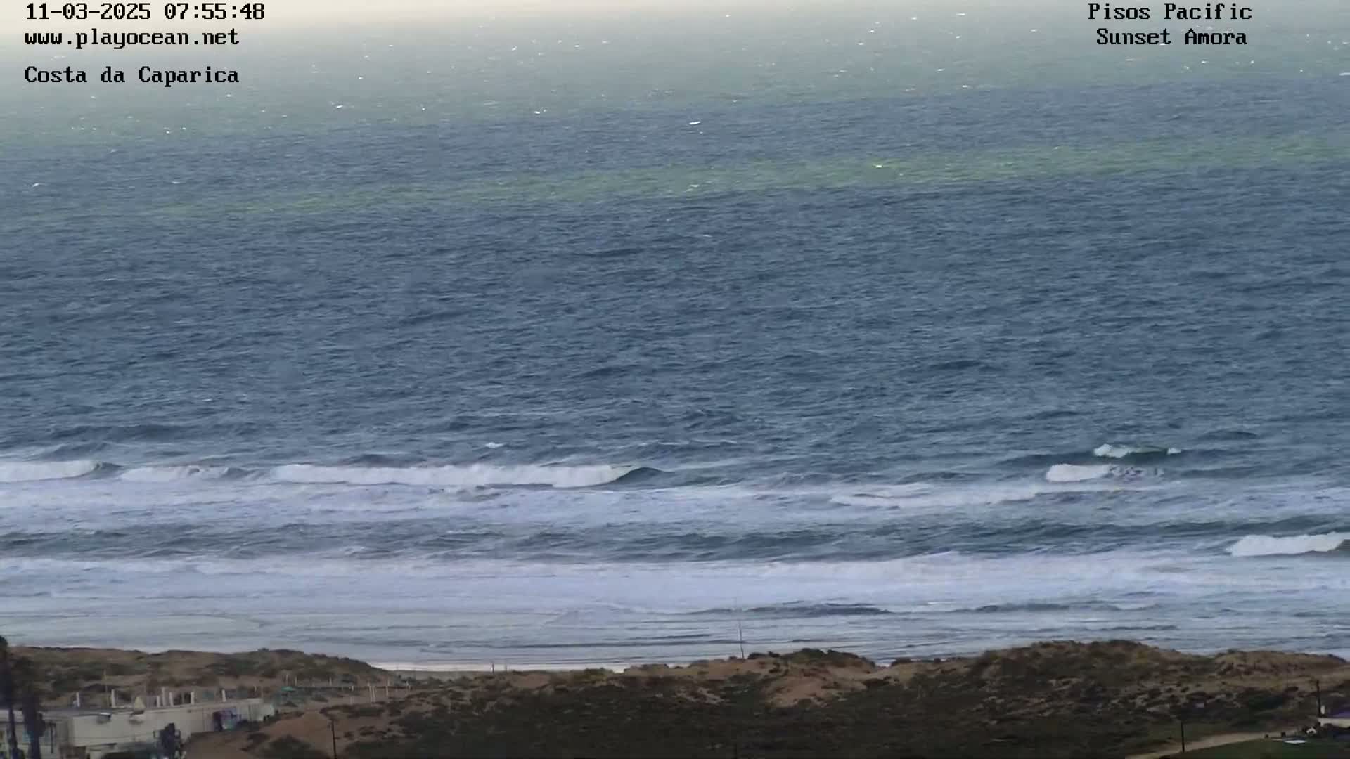 A hazy, overcast day shows a wide expanse of dark blue ocean with small waves breaking on a sandy shore, where some buildings are visible.