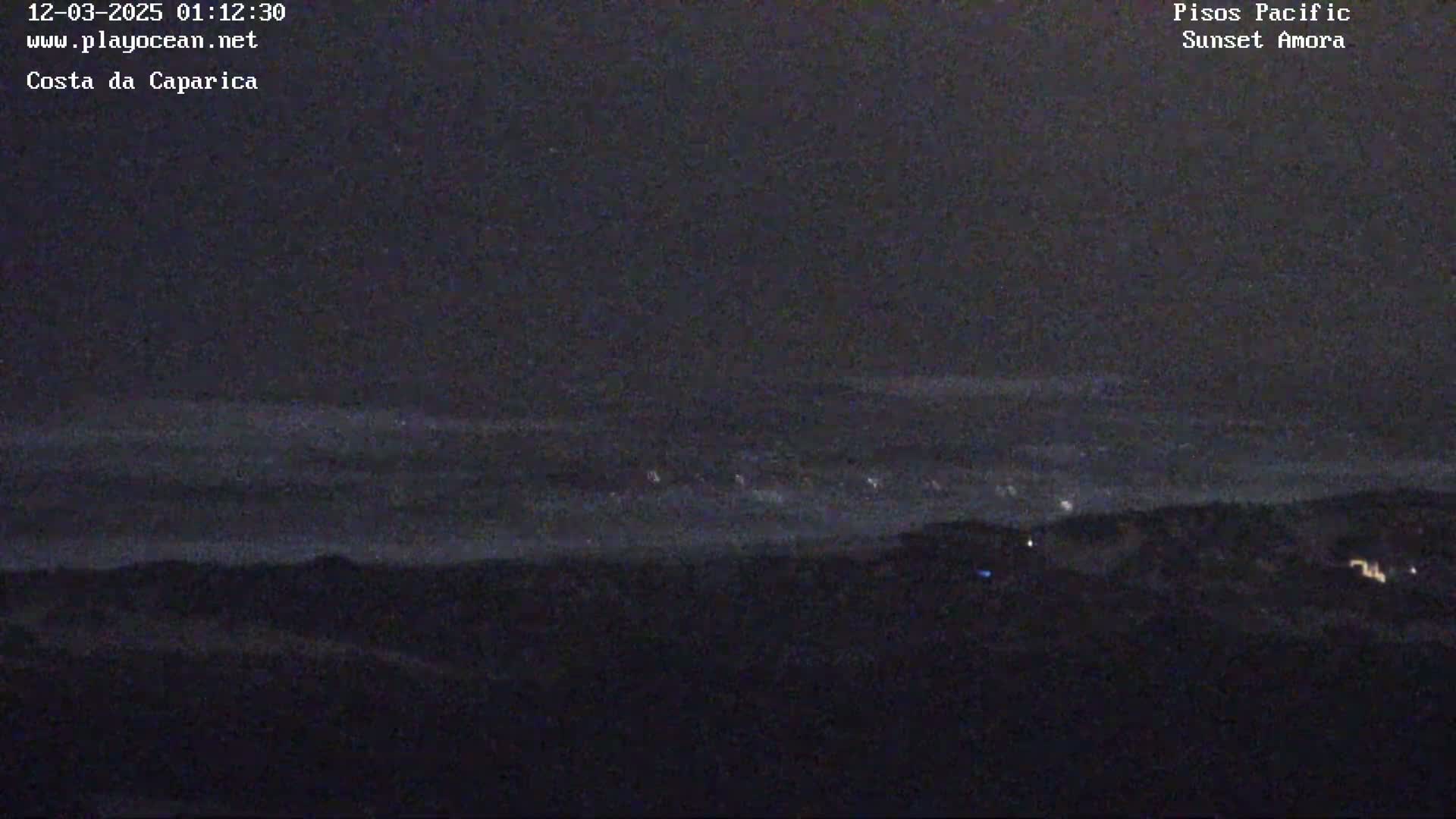 A dark, nighttime landscape shows a distant line of lights over a dark body of water under a mostly clear, dark sky.