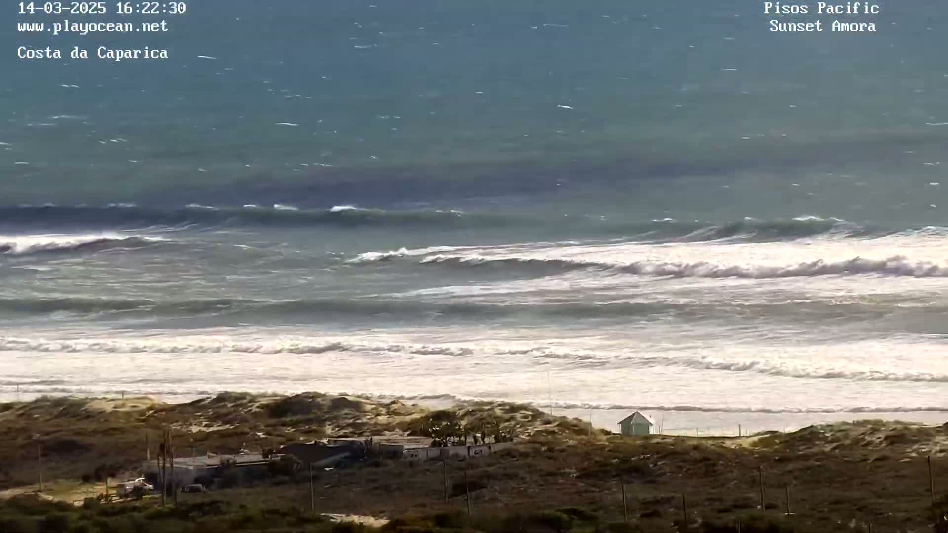 A high-angle, long shot shows a wavy ocean under a somewhat cloudy sky, with sandy dunes and a few buildings visible in the foreground.