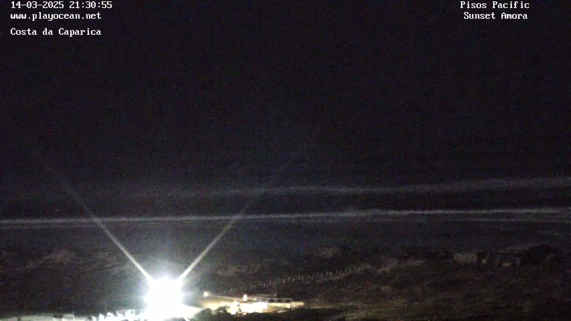 A nighttime coastal scene shows a dark ocean with faint waves under a clear, dark sky, illuminated by bright lights on the shore.