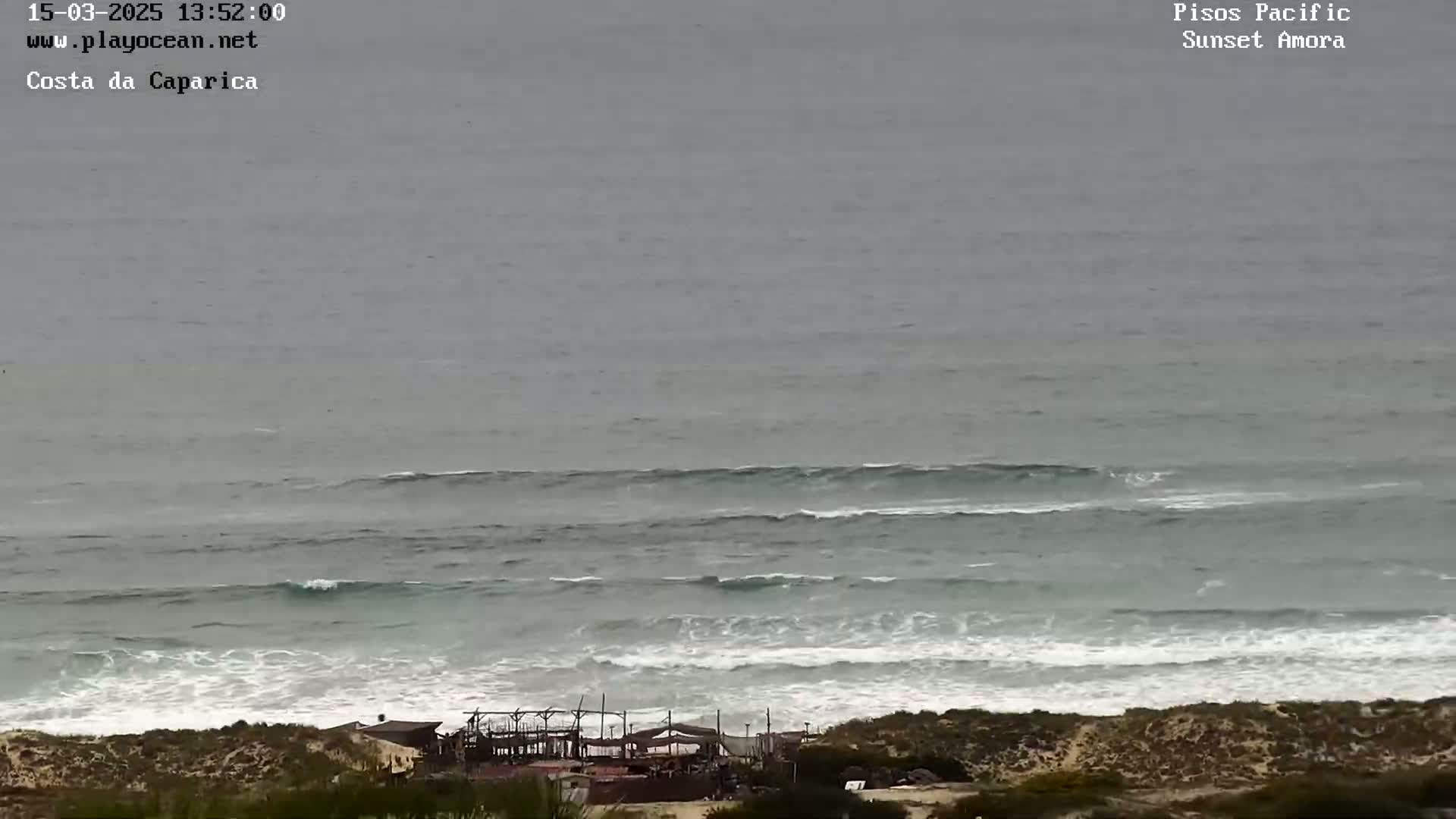 A high-angle, long shot shows a beach with moderate waves under an overcast sky, and a structure is visible near the shoreline.