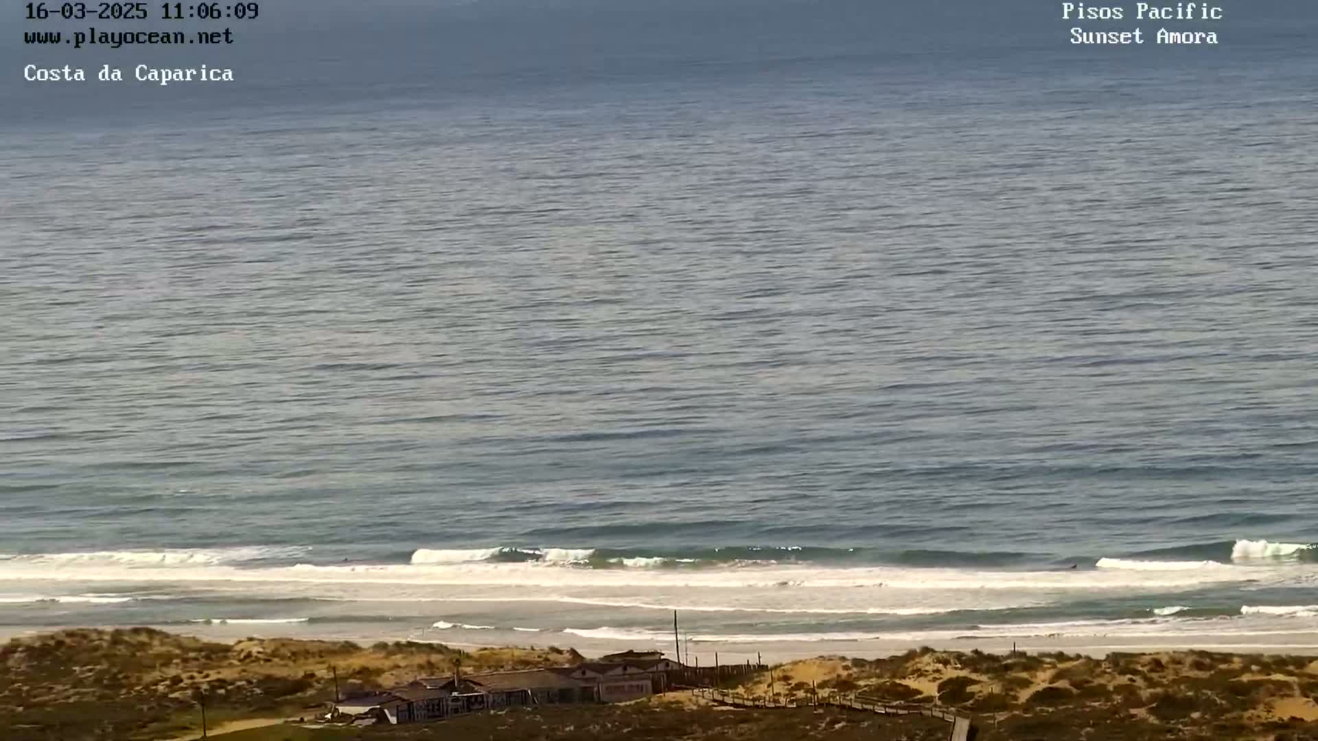 A long sandy beach with gentle waves and a few buildings is visible from an elevated vantage point on a clear day.