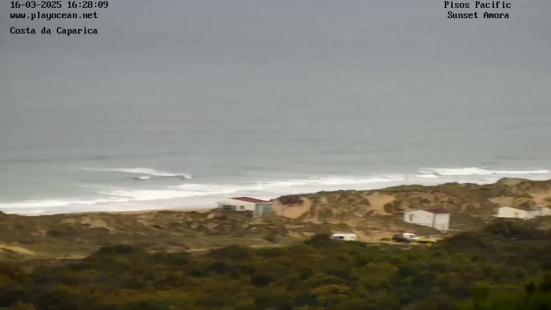 A high-angle view shows a gray, overcast day at a beach with several small houses nestled into the sand dunes, overlooking an ocean with small waves.