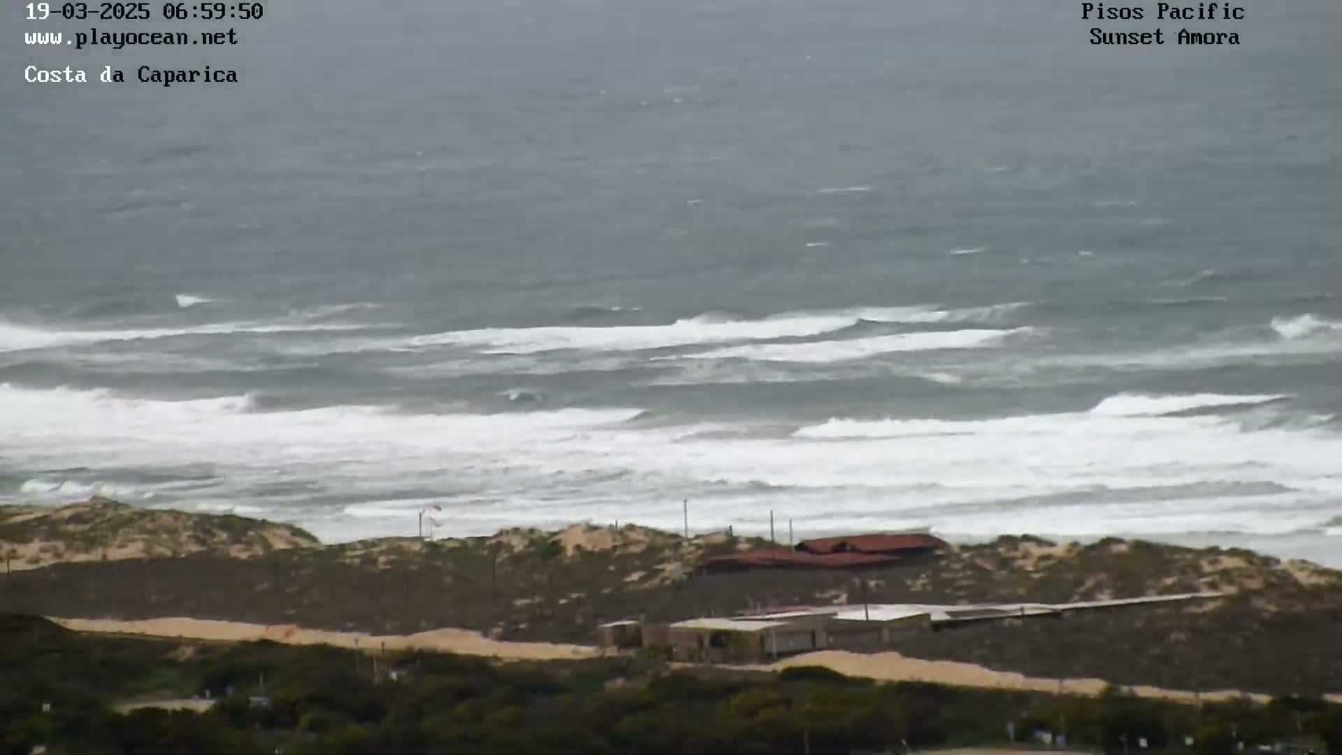A coastal scene shows a choppy ocean with white-capped waves breaking near a sandy shore with low-lying buildings and vegetation under an overcast sky.
