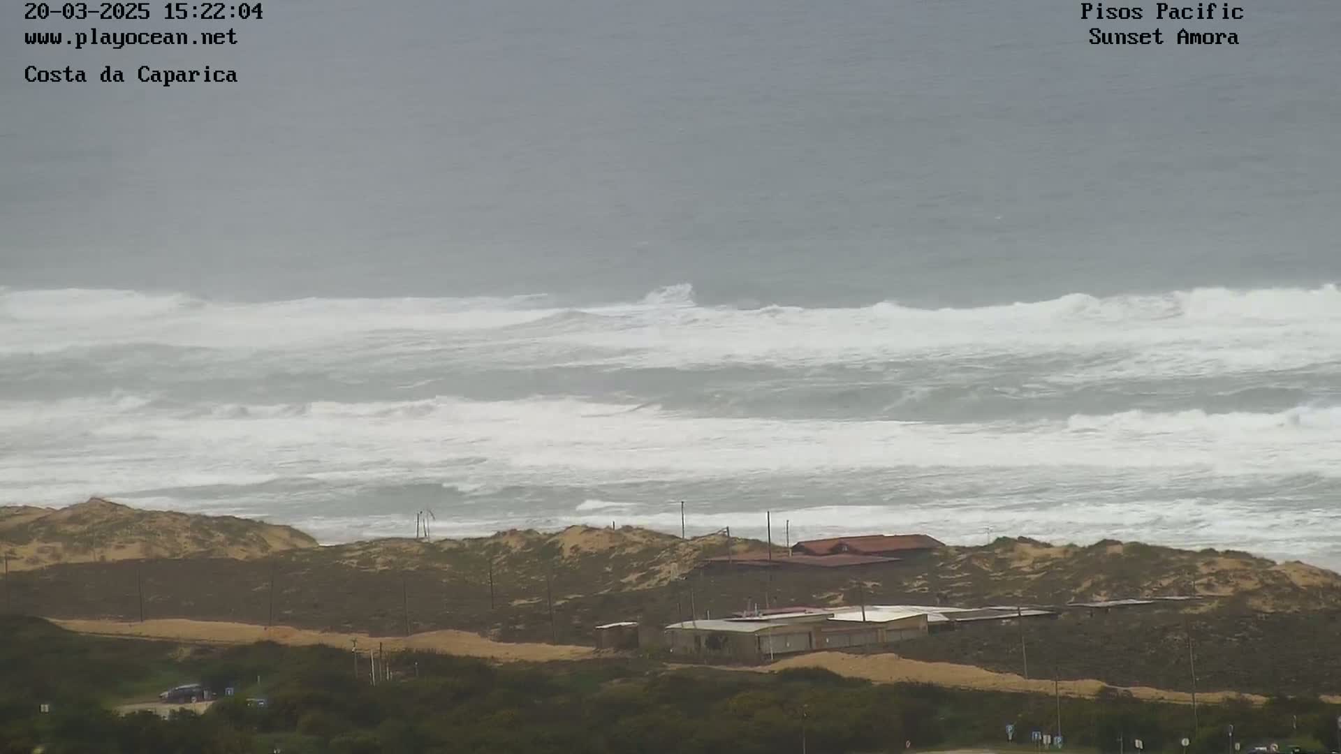Large, choppy waves crash on a beach with a few buildings and sandy dunes visible under an overcast sky.