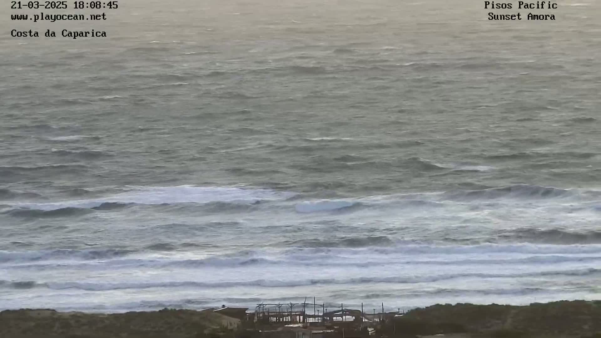A long shot of a choppy sea with moderate waves breaking near a shoreline with a few structures visible in the distance.