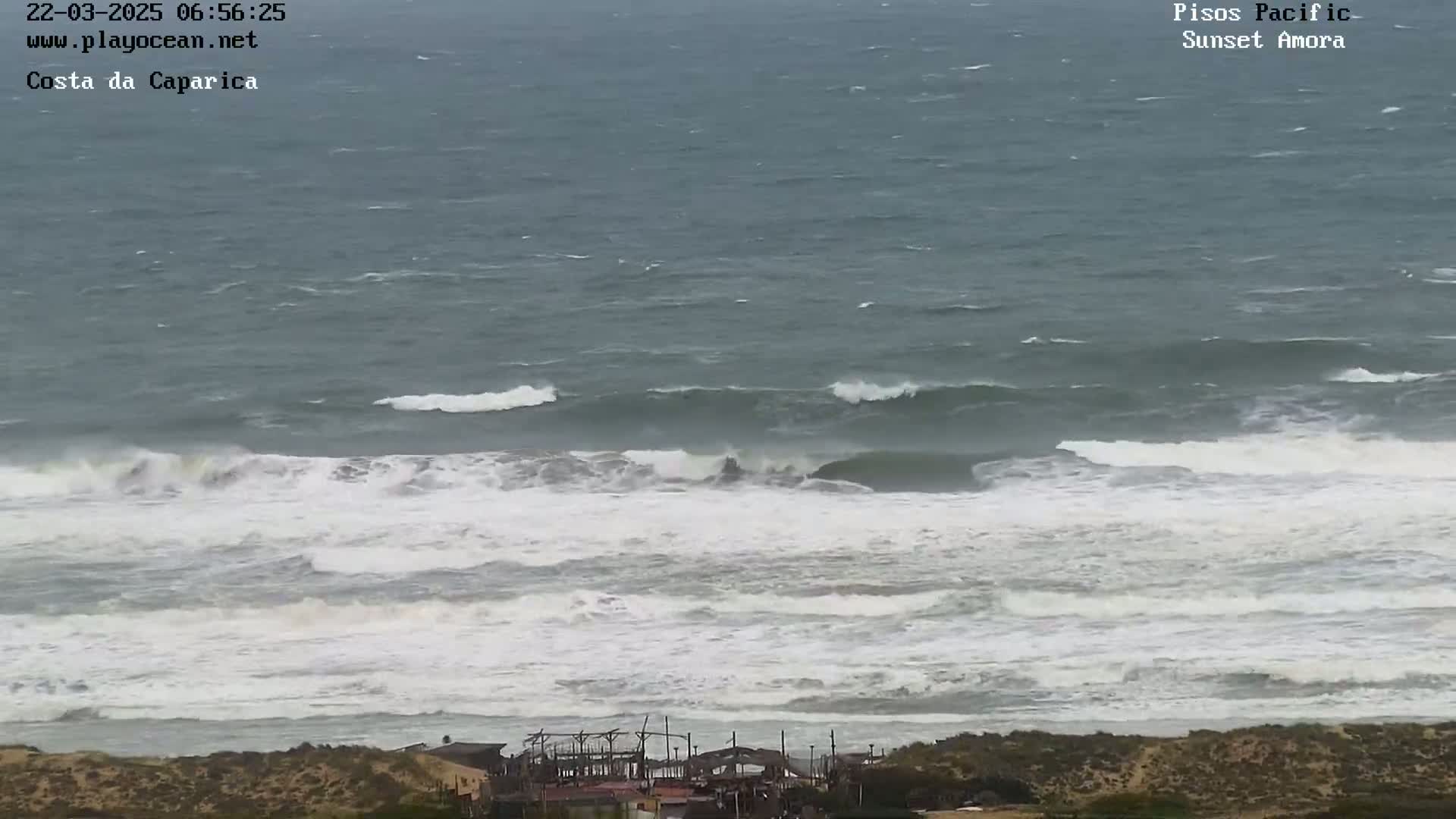 A long shot of a stormy ocean with large, white-capped waves crashing near a sandy shoreline with some structures visible in the distance.