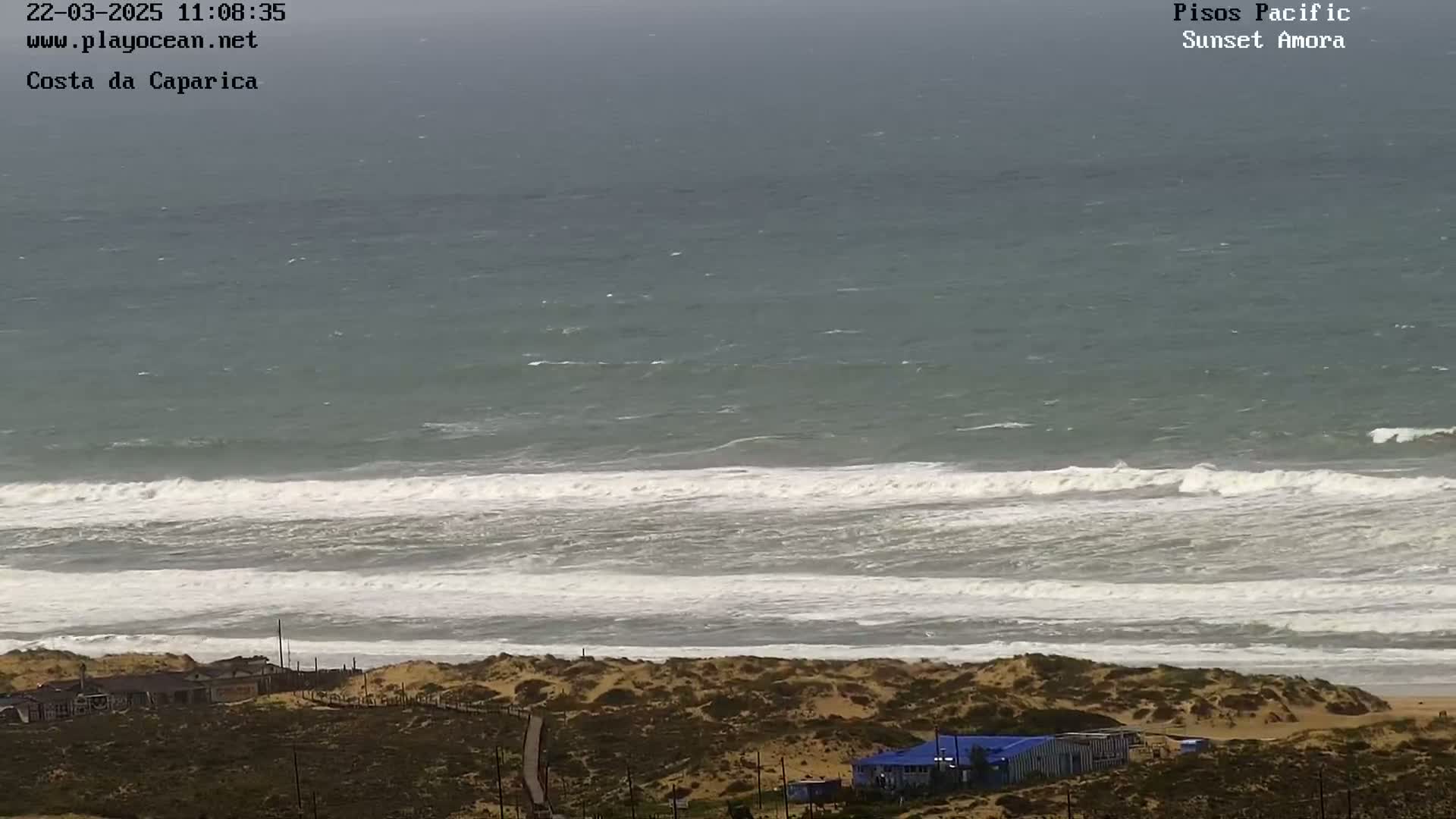 A windy day shows a wide expanse of ocean with white-capped waves rolling onto a sandy beach with buildings visible in the distance.