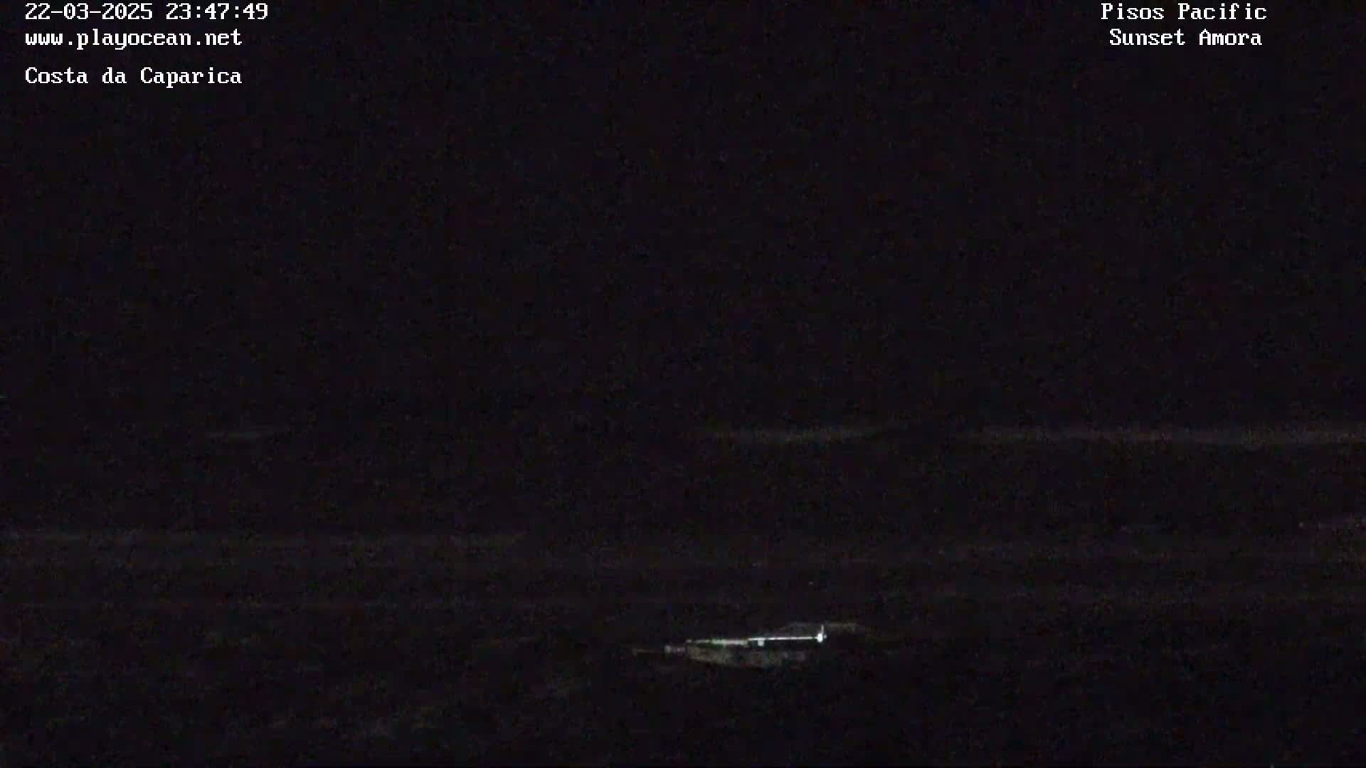 A nighttime view of a dark ocean with a faint horizon line and a few small lights visible in the distance.