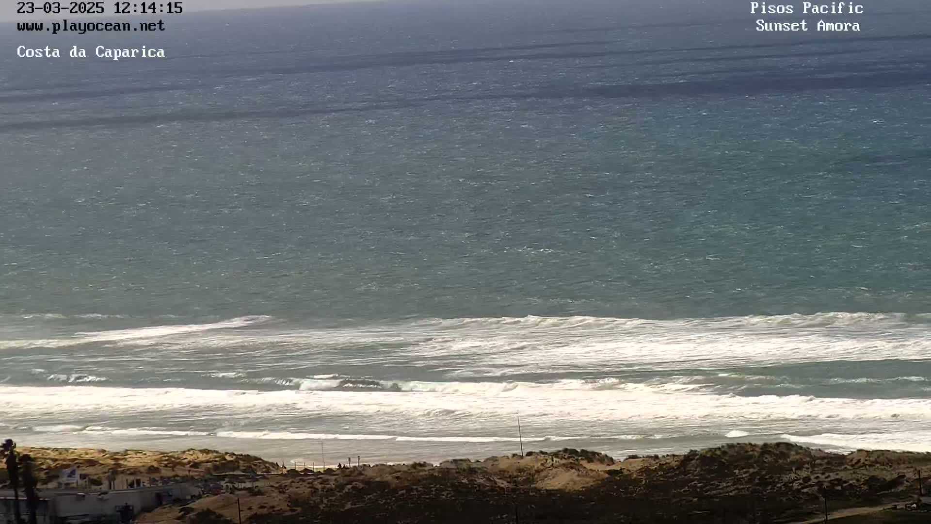 A high-angle, long shot shows a beach with waves breaking on the shore under a partly cloudy sky.