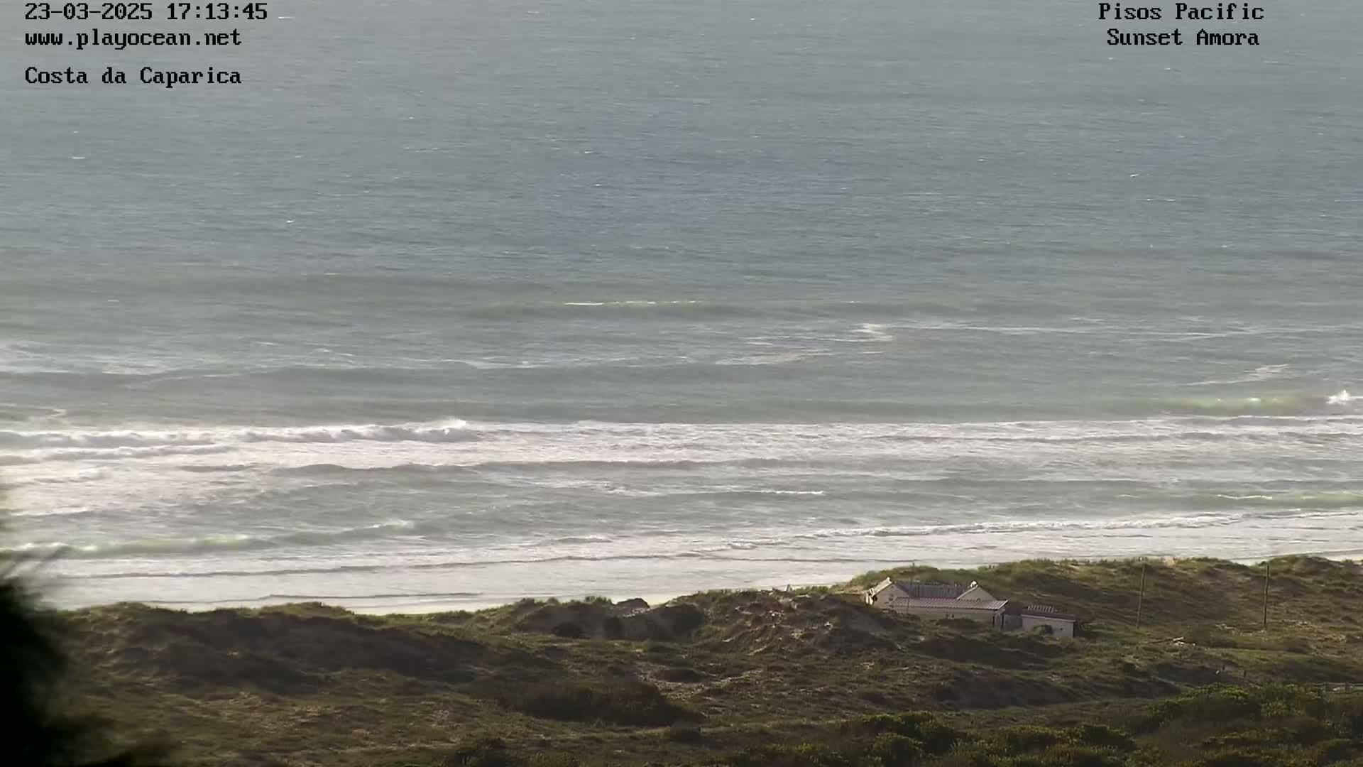 A coastal scene shows a calm ocean with small waves breaking on a sandy beach, behind which sits a small building nestled in dune vegetation.