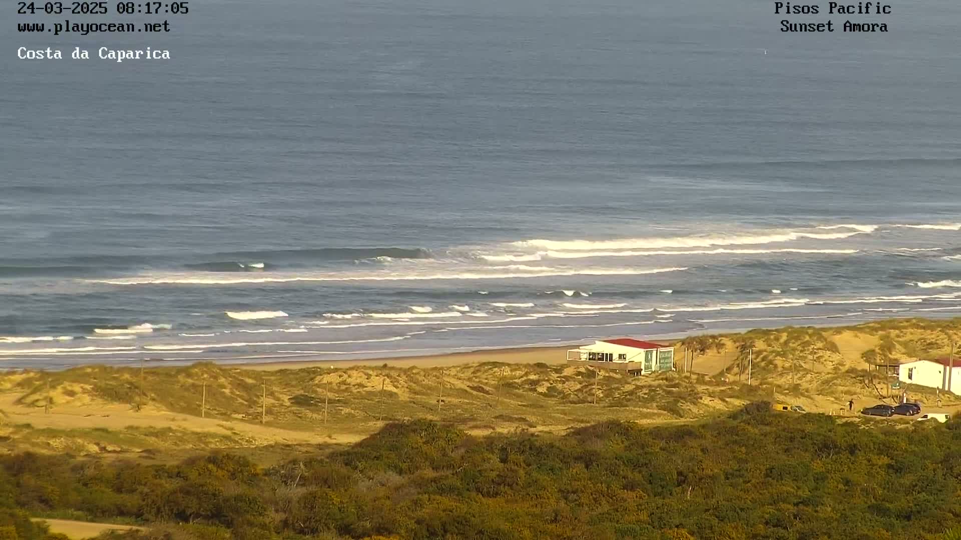 A beach with rolling waves under a mostly clear sky, showing a sandy shoreline with dune grasses and a few buildings.