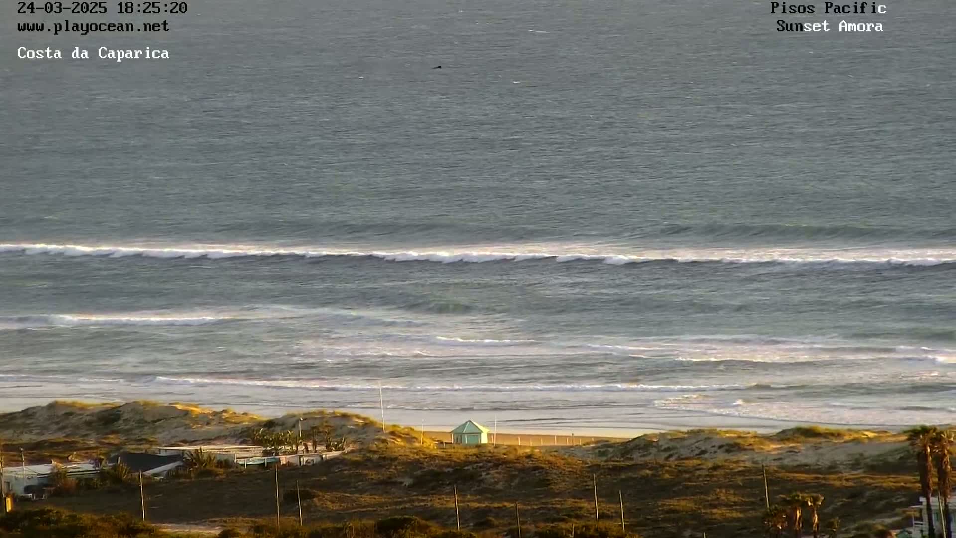 A long expanse of ocean with waves rolling onto a sandy beach, backed by low-lying vegetation and a small building, under a partly sunny sky.
