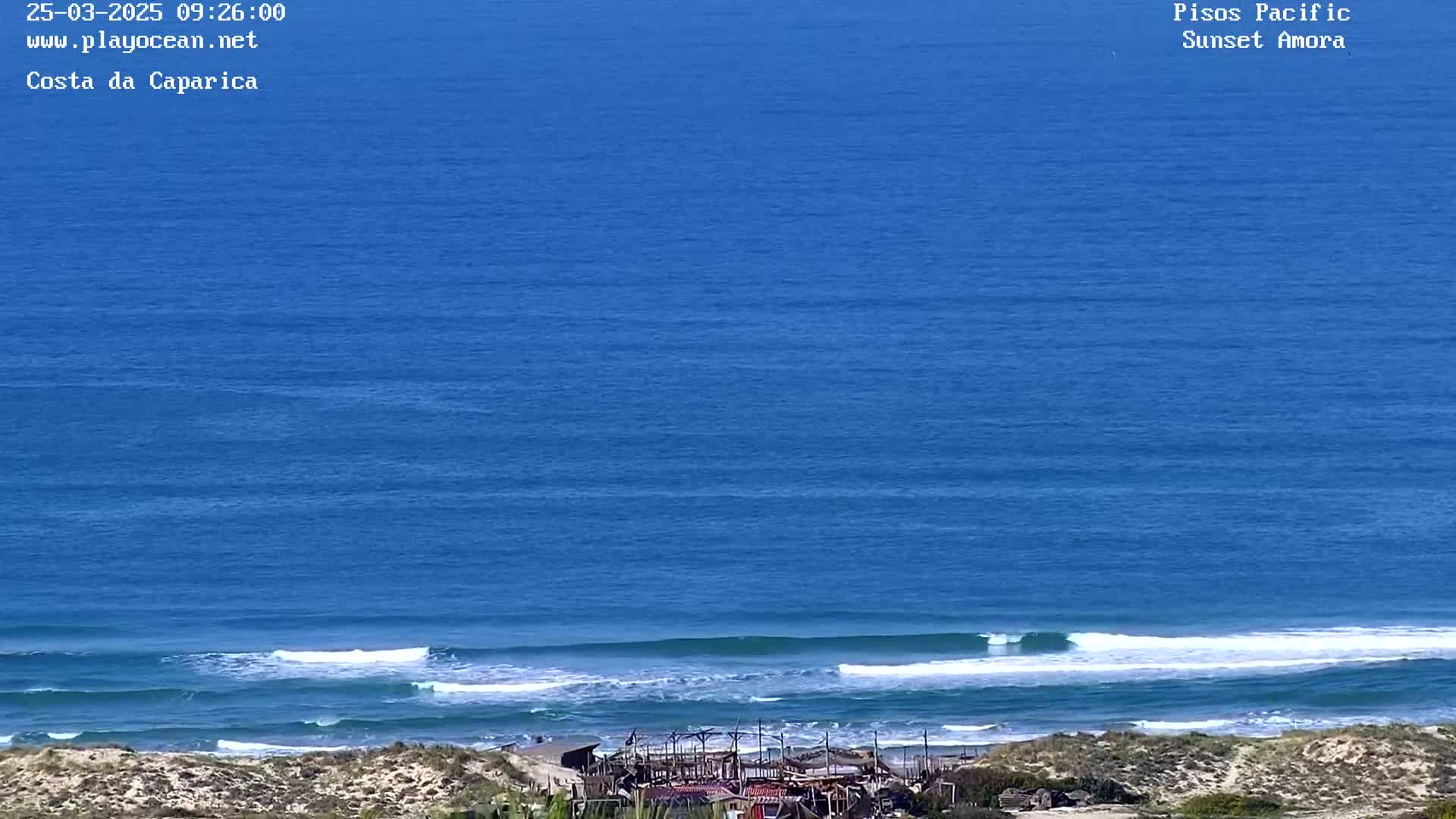A high-angle view shows a calm ocean with small waves breaking on a sandy shore under a clear blue sky.