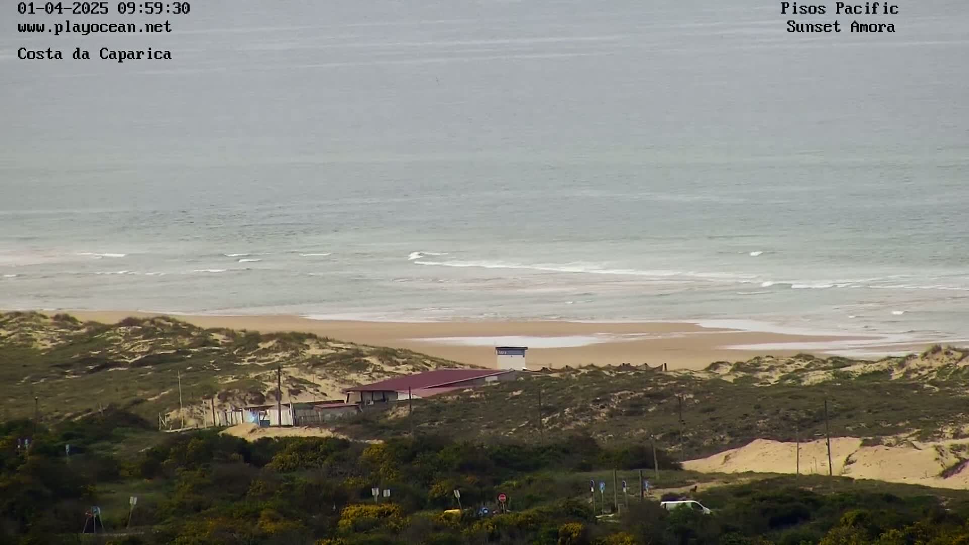 A beach with calm waves and a building behind sand dunes is viewed from a distance on an overcast day.