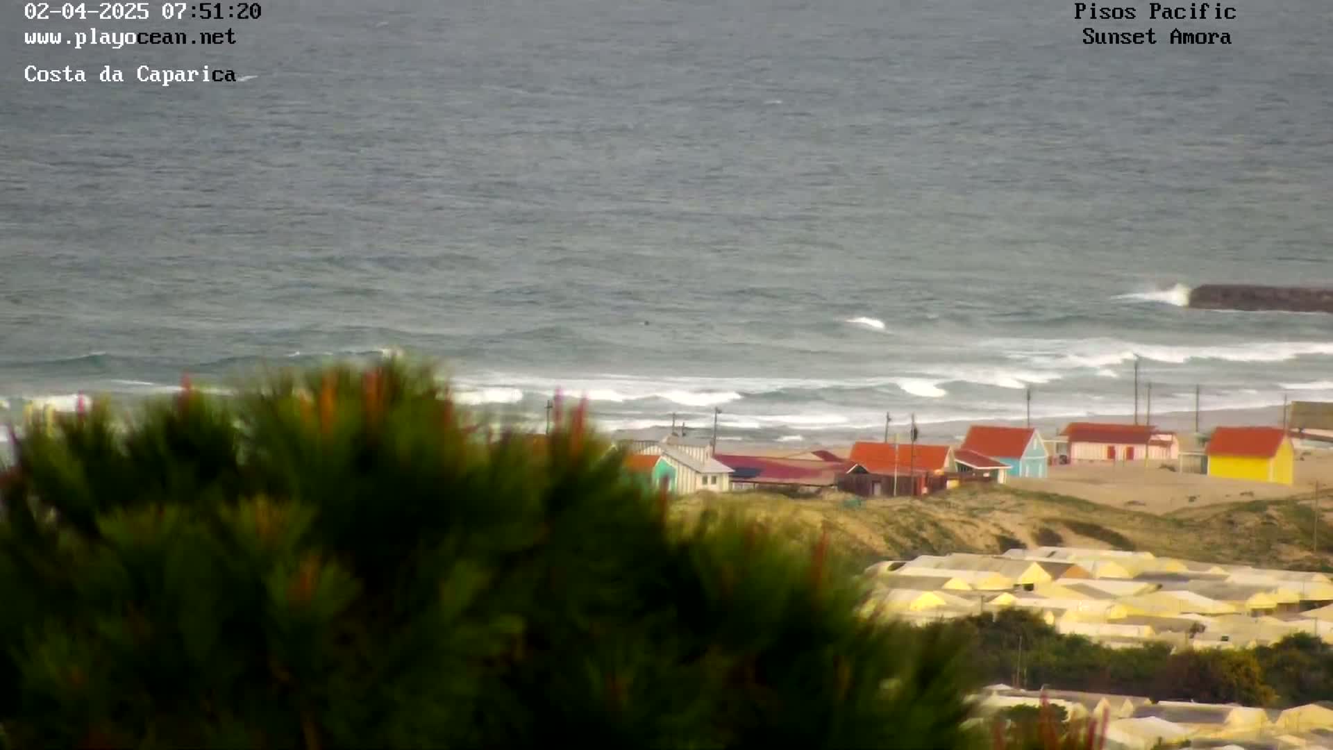 A coastal view shows a gray, somewhat choppy ocean with waves breaking on a sandy beach, behind which are rows of small, colorful houses, partially obscured by green vegetation in the foreground.