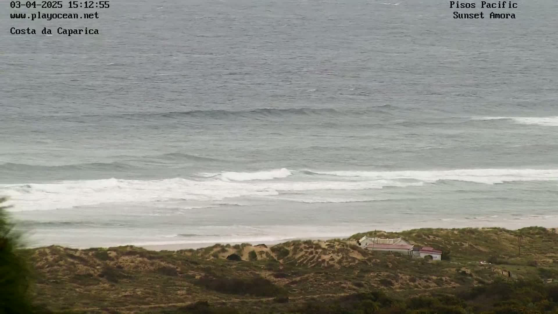 A high-angle view shows a sandy beach with a building, backed by low-lying vegetation, and a grey ocean with white-capped waves rolling toward the shore.