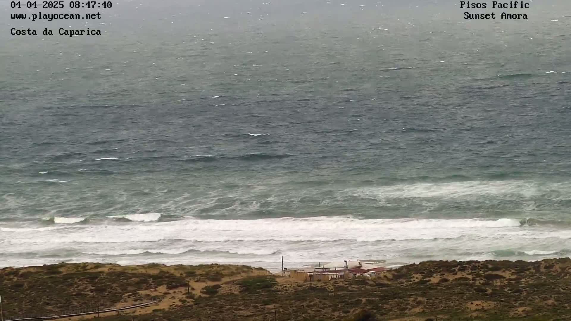 A high-angle view shows a beach with white-capped waves crashing on the shore under an overcast sky, and a small building is visible on the dunes.