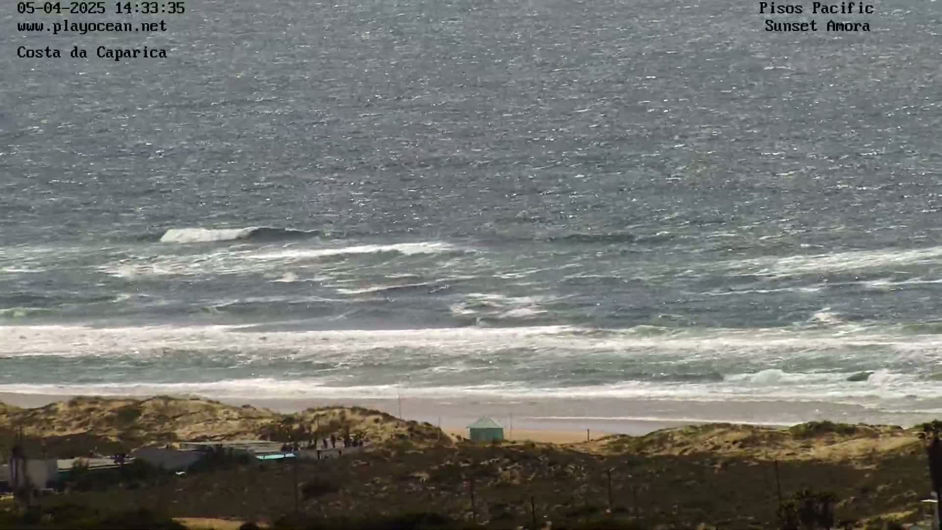 A view from a hilltop shows a sandy beach with a small building and some people, and a choppy ocean with waves breaking near the shore under a partly cloudy sky.