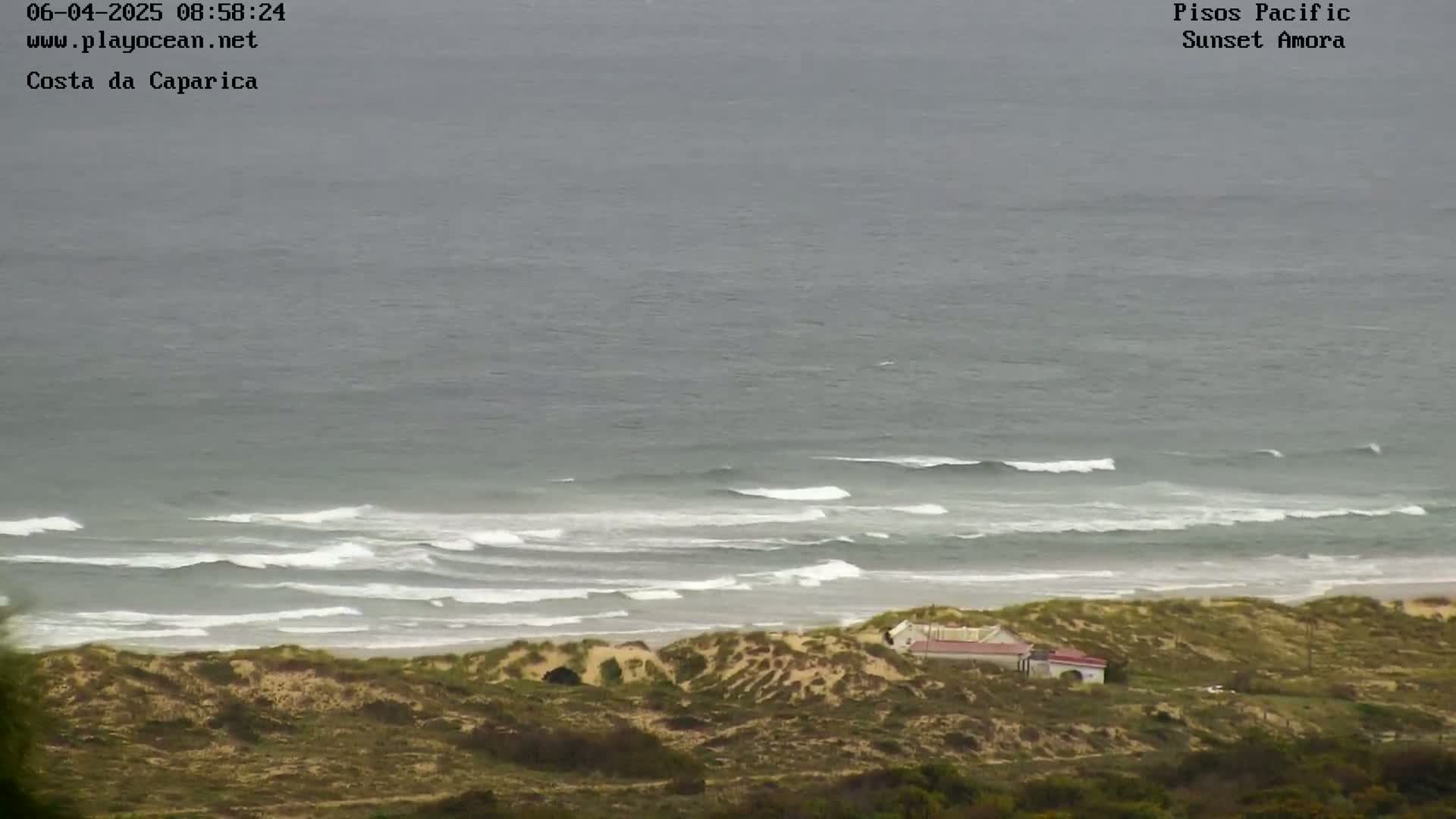 A high-angle view shows a gray, overcast day at a beach with moderate waves breaking near the shore, and a small building nestled among sand dunes.