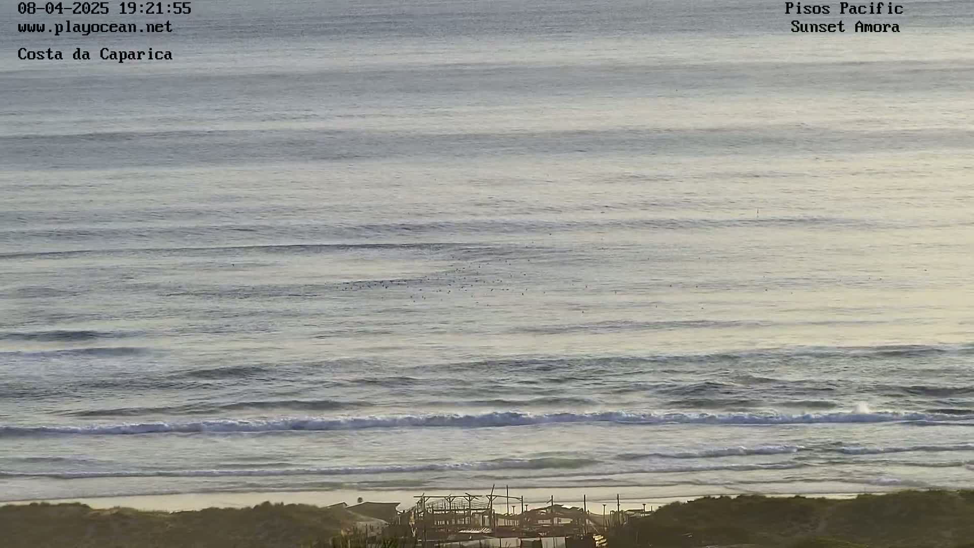 A long, wide shot of an ocean beach at sunset shows waves gently rolling toward the shore, with a small structure visible on the beach in the foreground.