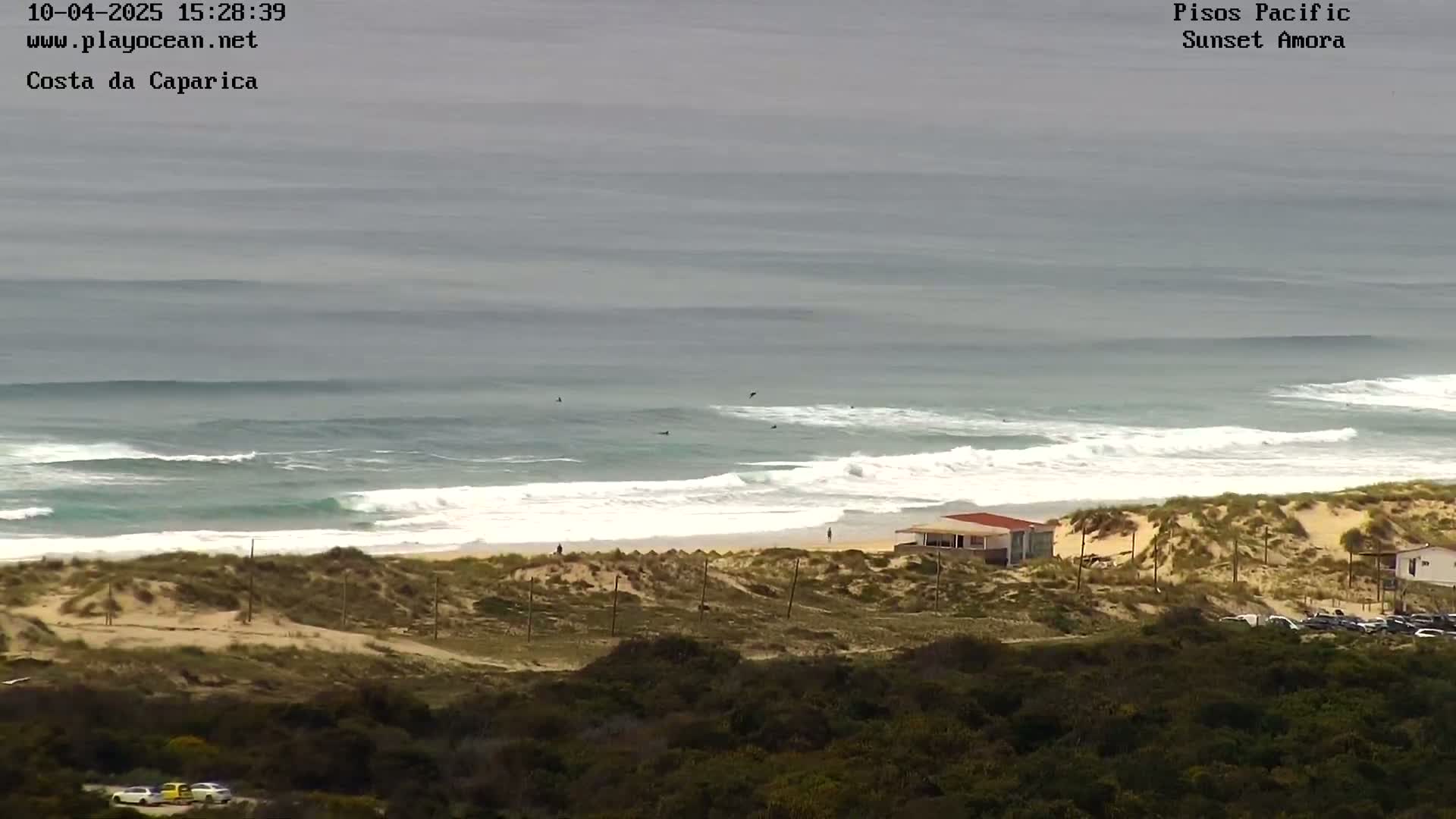 An ocean beach with several surfers in the water, sand dunes, and a few small buildings, under an overcast sky.