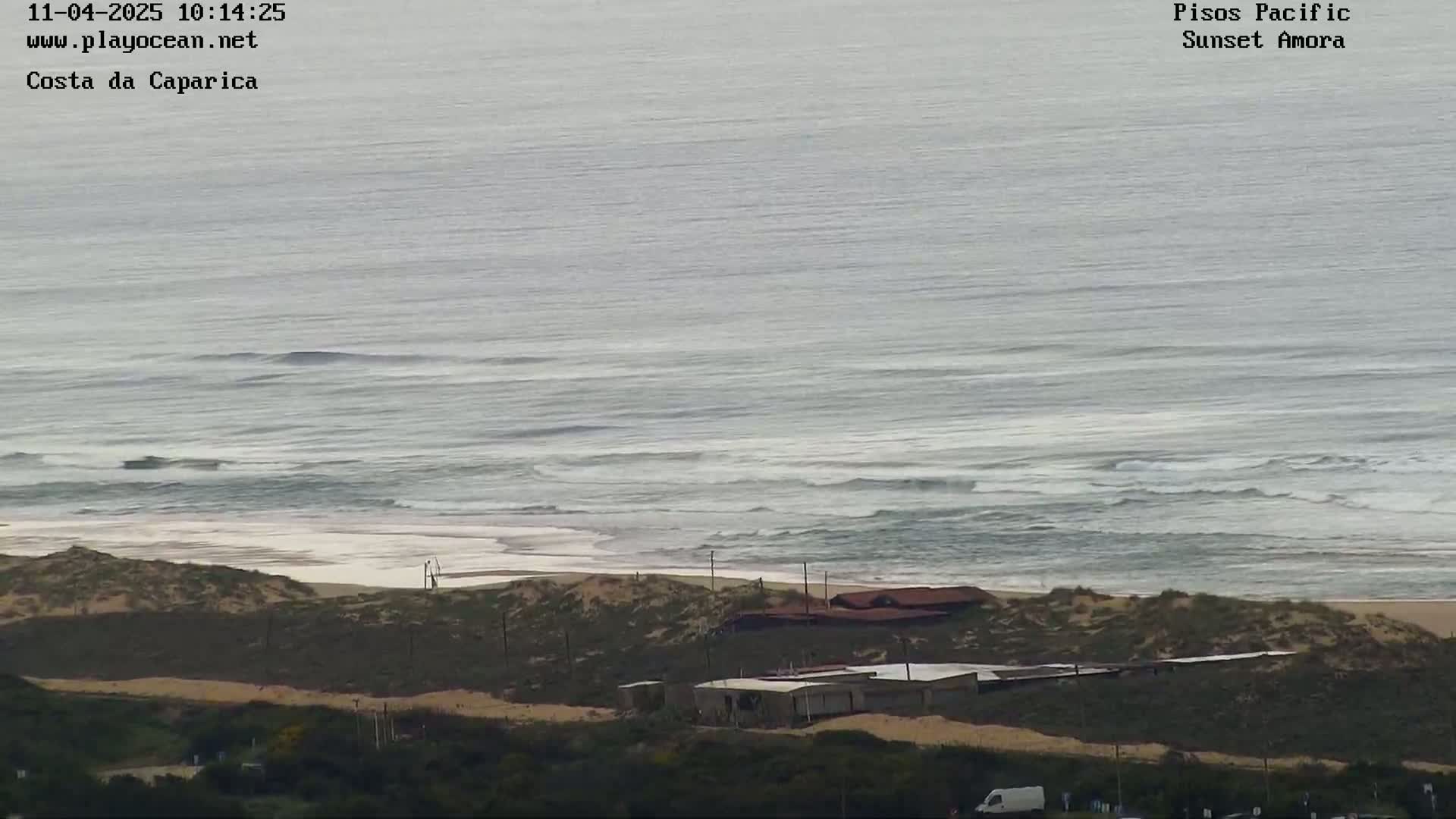 A high-angle view shows a sandy beach with some buildings behind it, overlooking a gray ocean with small waves under an overcast sky.