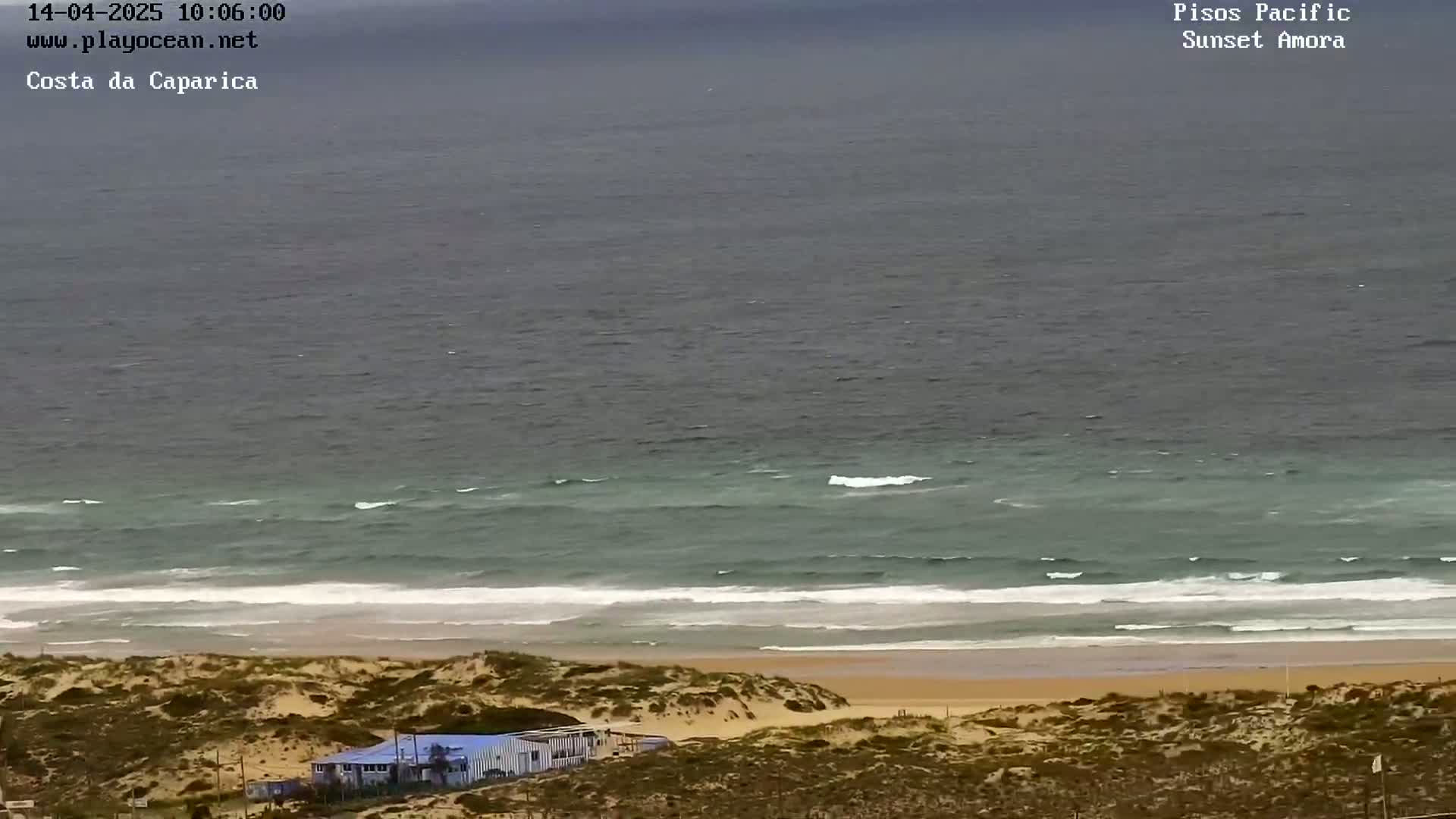 A sandy beach with a building and some vegetation overlooks a moderately wavy ocean under a cloudy sky.
