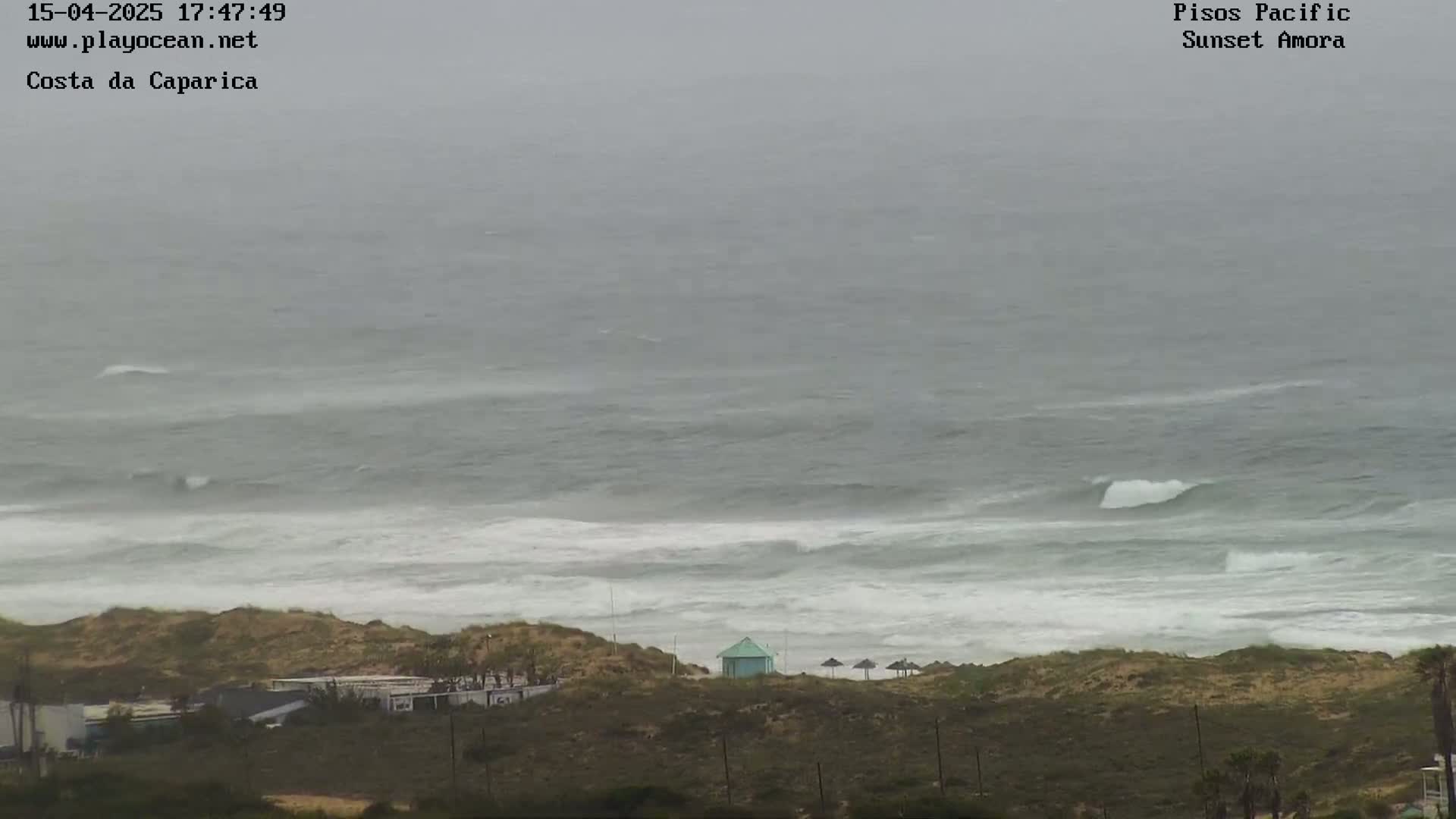 A hazy, overcast day shows a beach with a small teal building and a few umbrellas on the sand dunes overlooking a rough, gray ocean with white-capped waves.