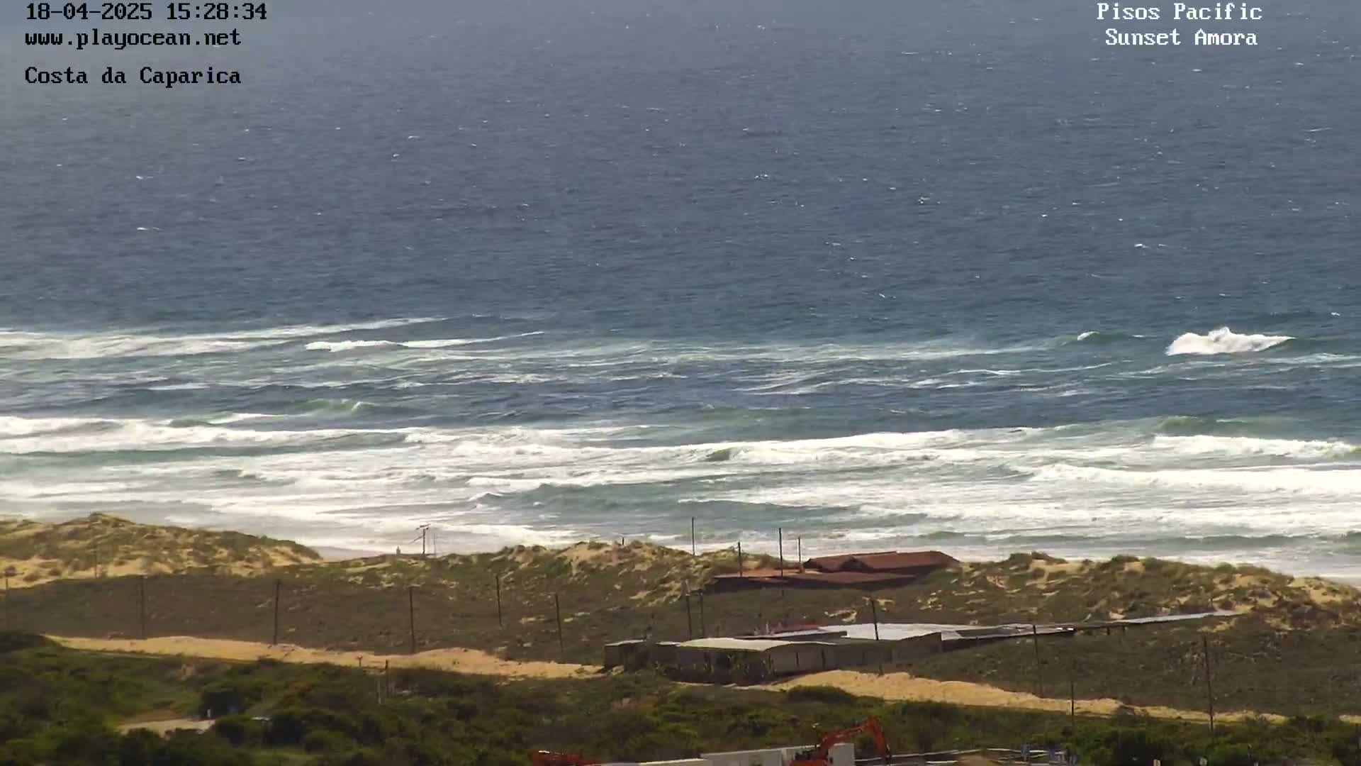 A high-angle view shows a sandy beach with some buildings and scrubby vegetation, overlooking a dark blue ocean with white-capped waves under a mostly clear sky.