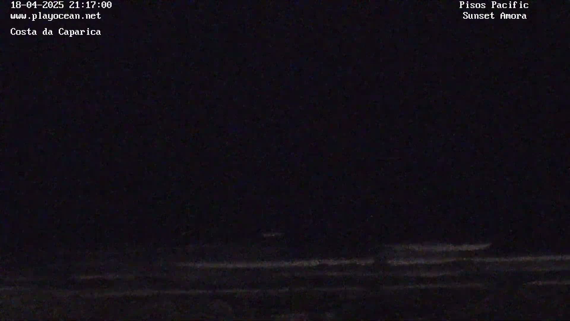 A dark nighttime ocean scene shows a shoreline with subtle wave action under a mostly clear, dark sky.