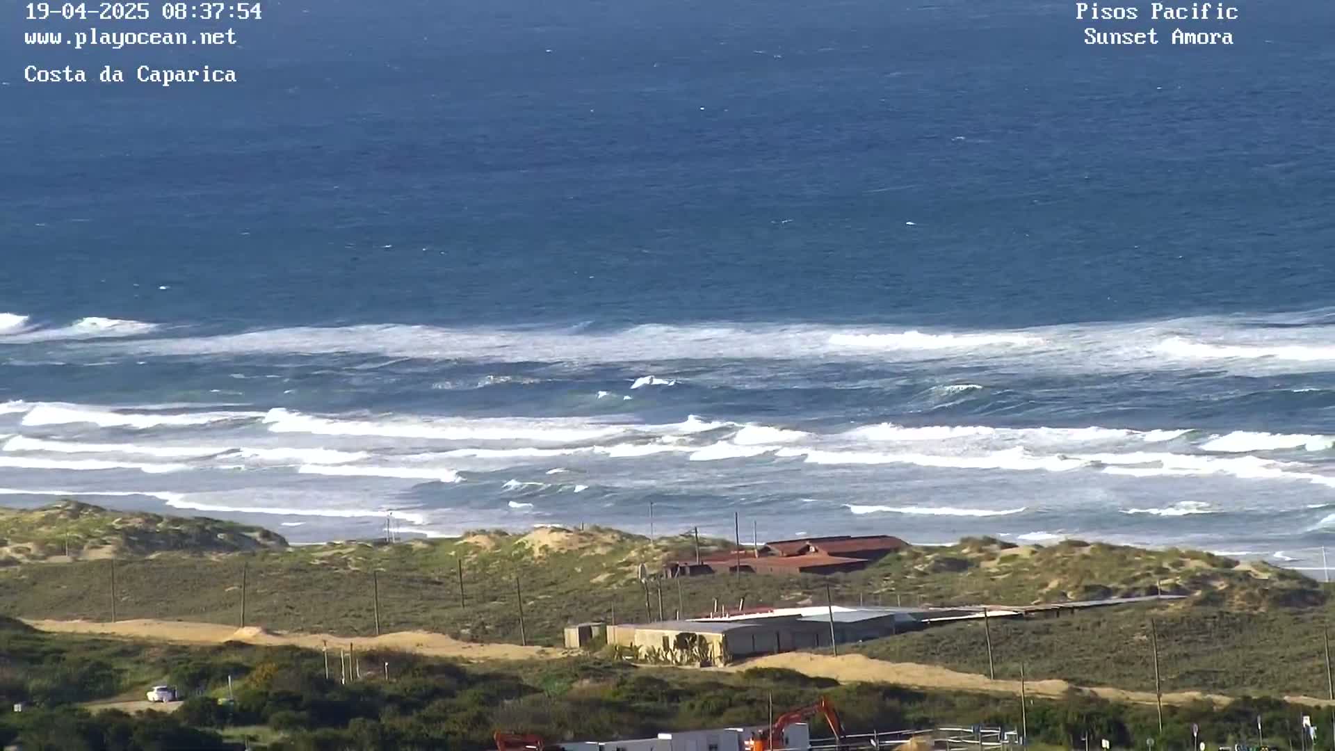 A sandy coastline with a few buildings is shown, with a vast ocean featuring rolling waves under a clear sky.