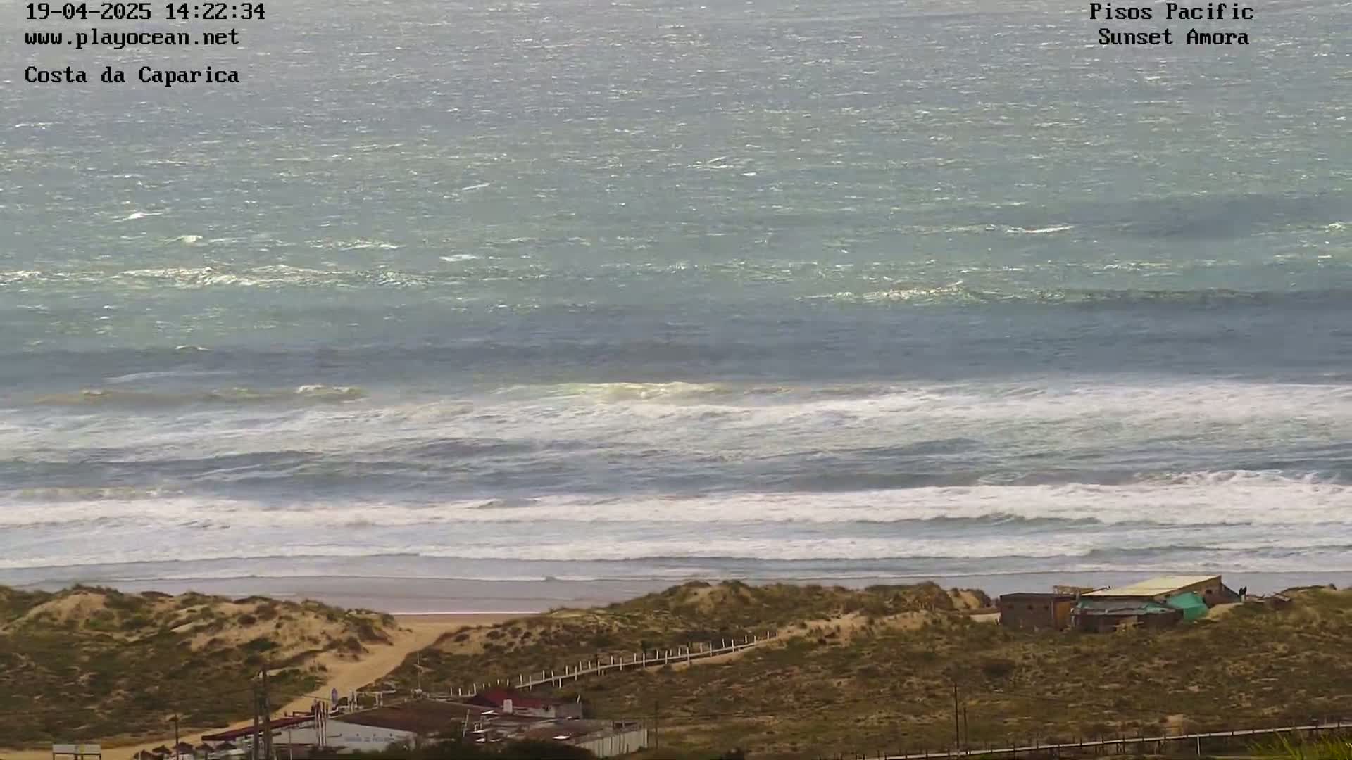 A windy day shows a choppy ocean meeting a sandy beach with dunes and a few buildings behind it.