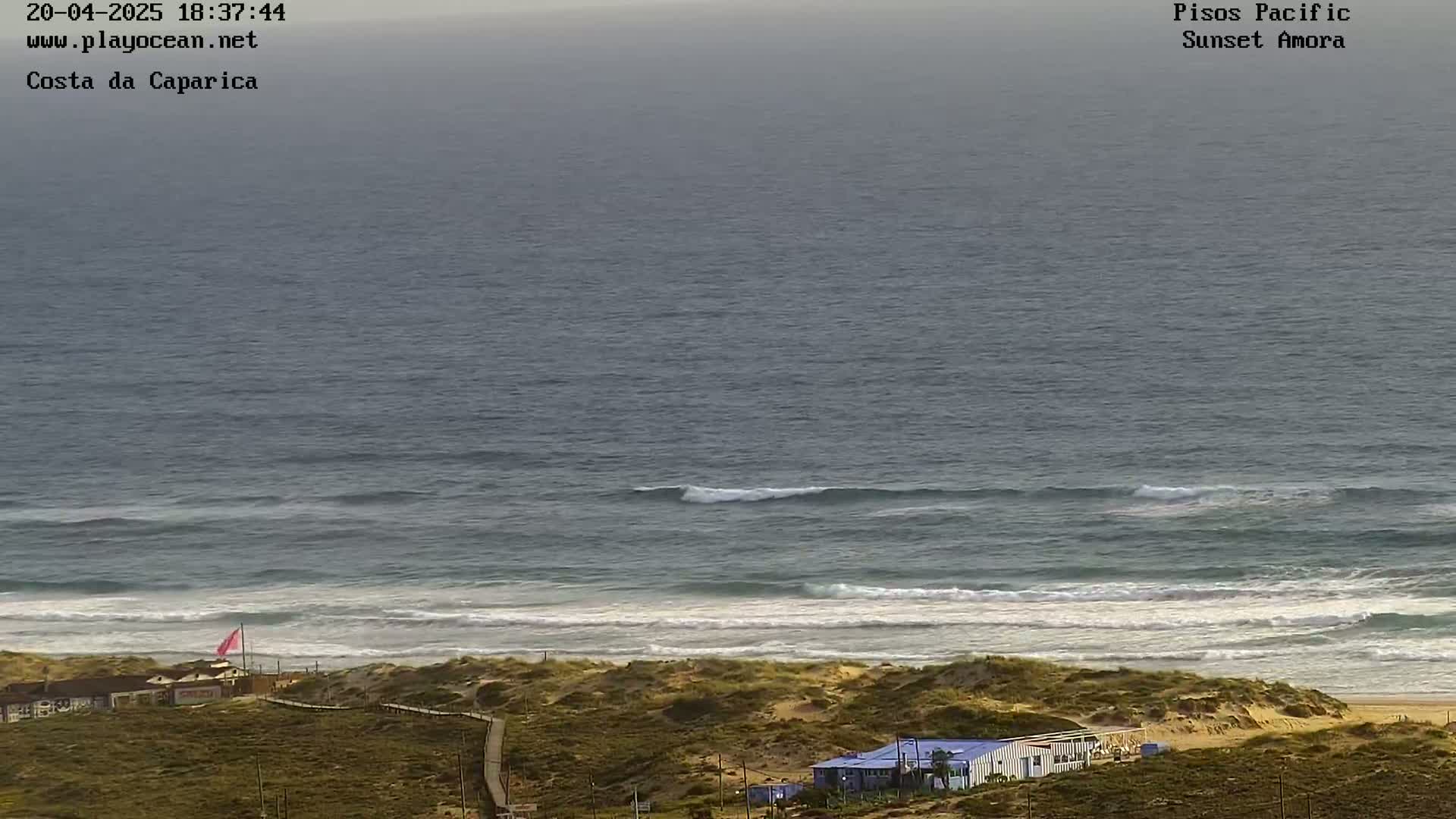 A beach with calm waves, sandy dunes, and a few buildings is seen under an overcast sky.