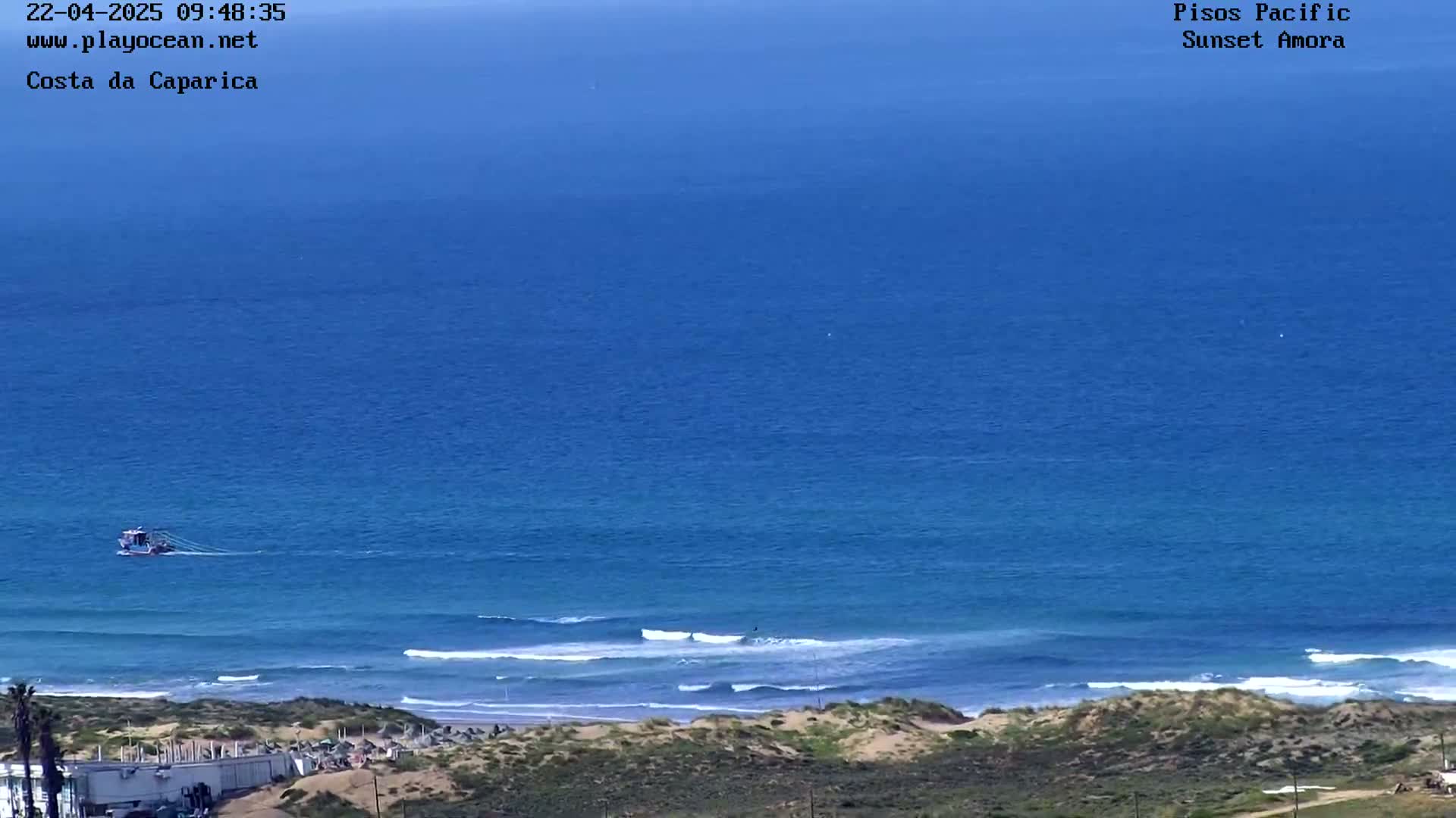 A small boat is visible on a calm, blue ocean with gentle waves breaking on a sandy shore.