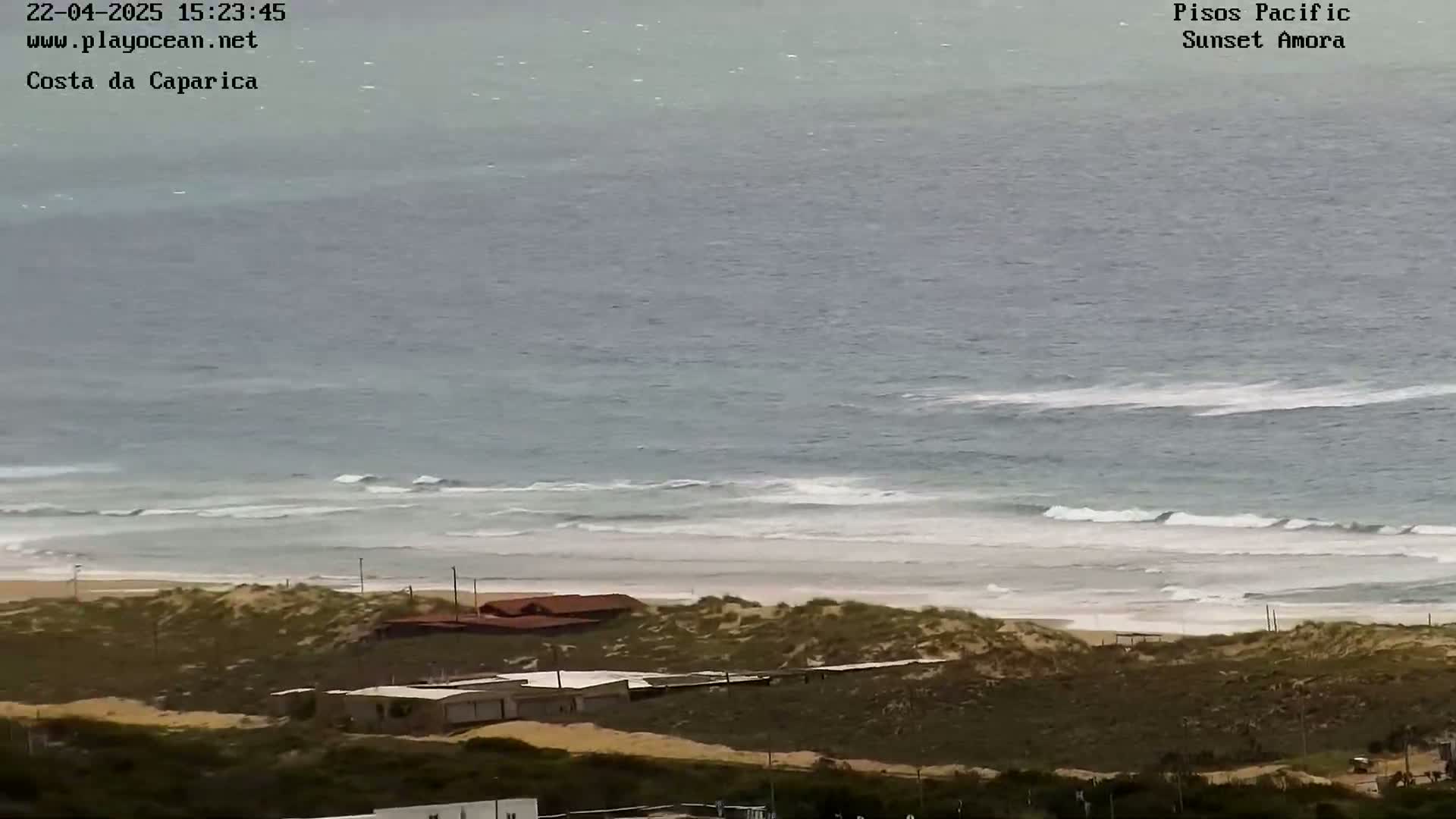 A coastal scene shows a sandy beach with low-lying buildings behind sand dunes and a somewhat choppy ocean with small waves under an overcast sky.