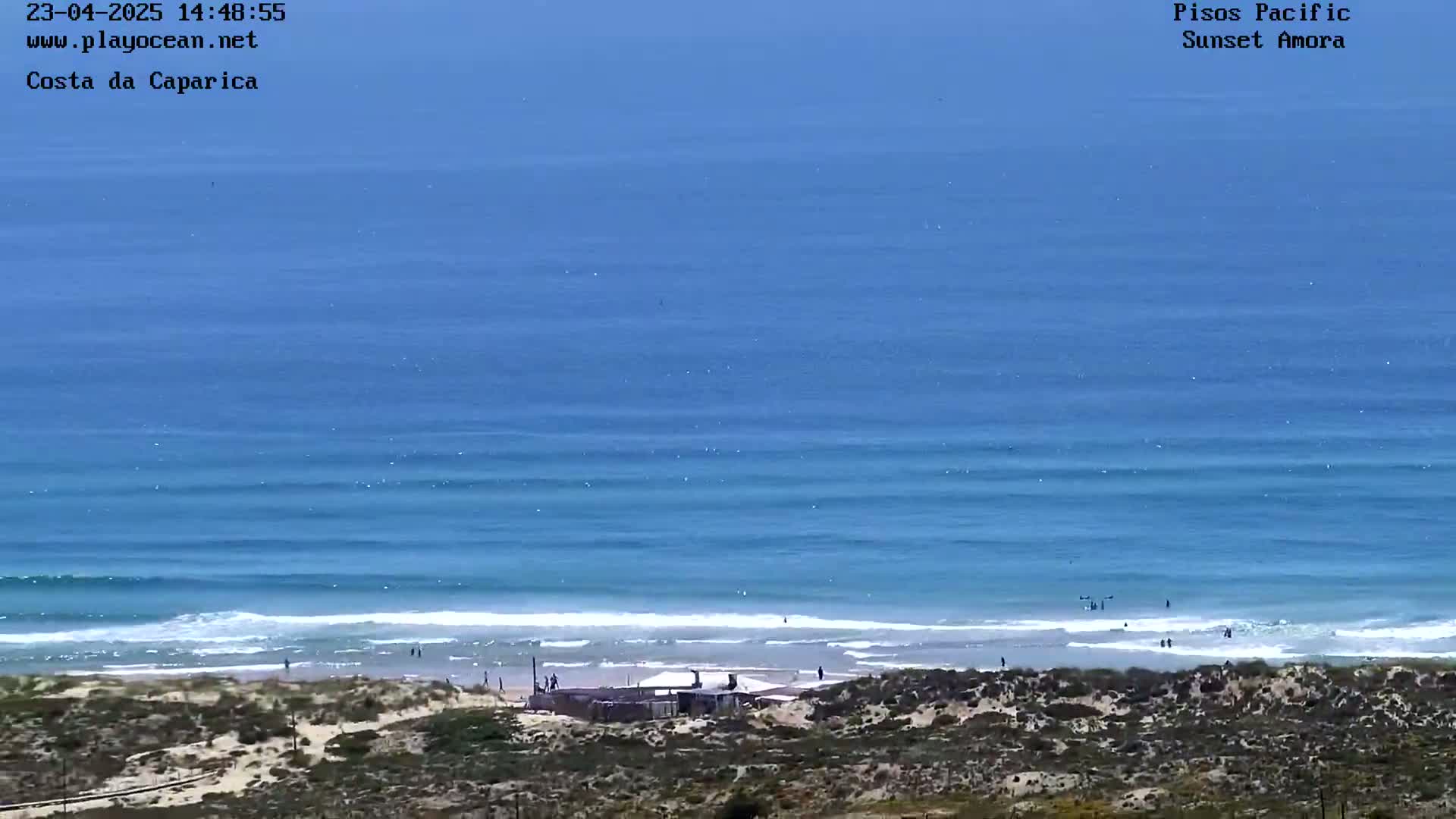 A wide shot shows a sandy beach with a small building and a few people, leading to a calm ocean with gentle waves under a clear, sunny sky.