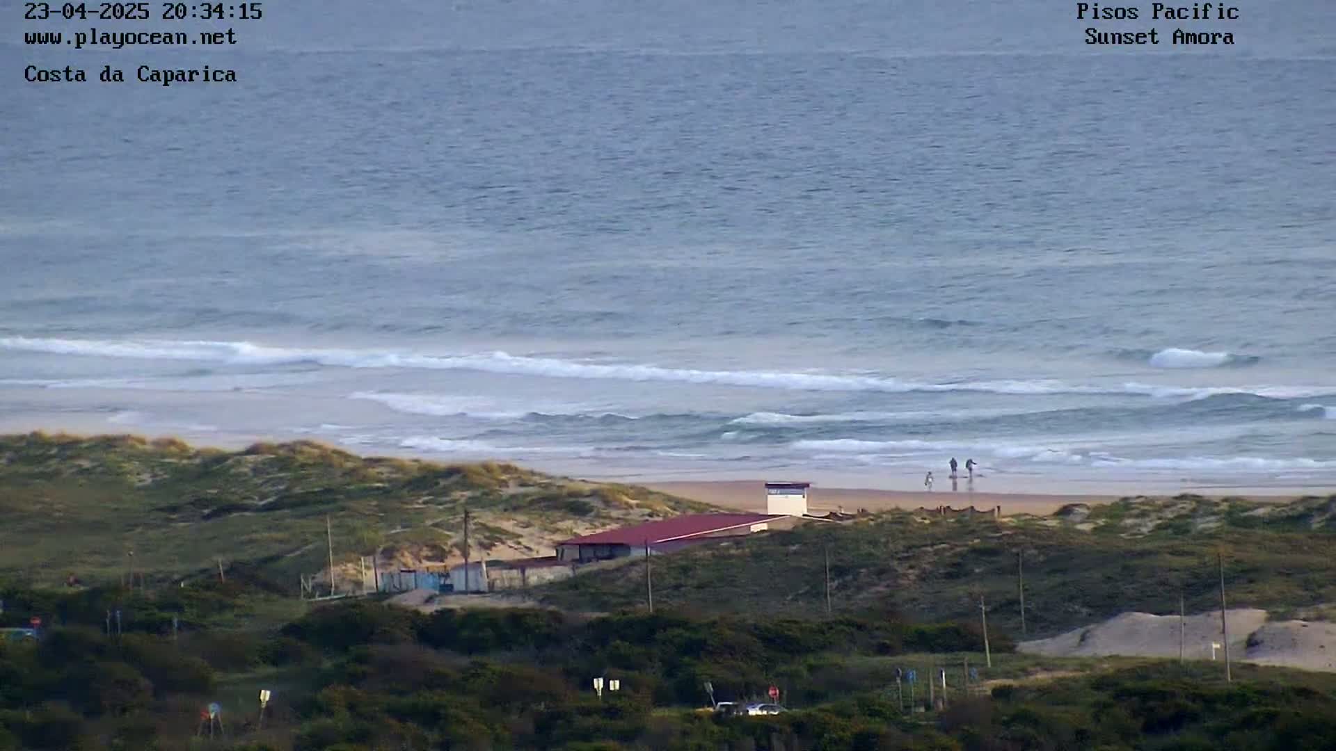 A long expanse of beach with gentle waves and a few people walking along the shoreline is overlooked by a low-lying building nestled among green dunes.
