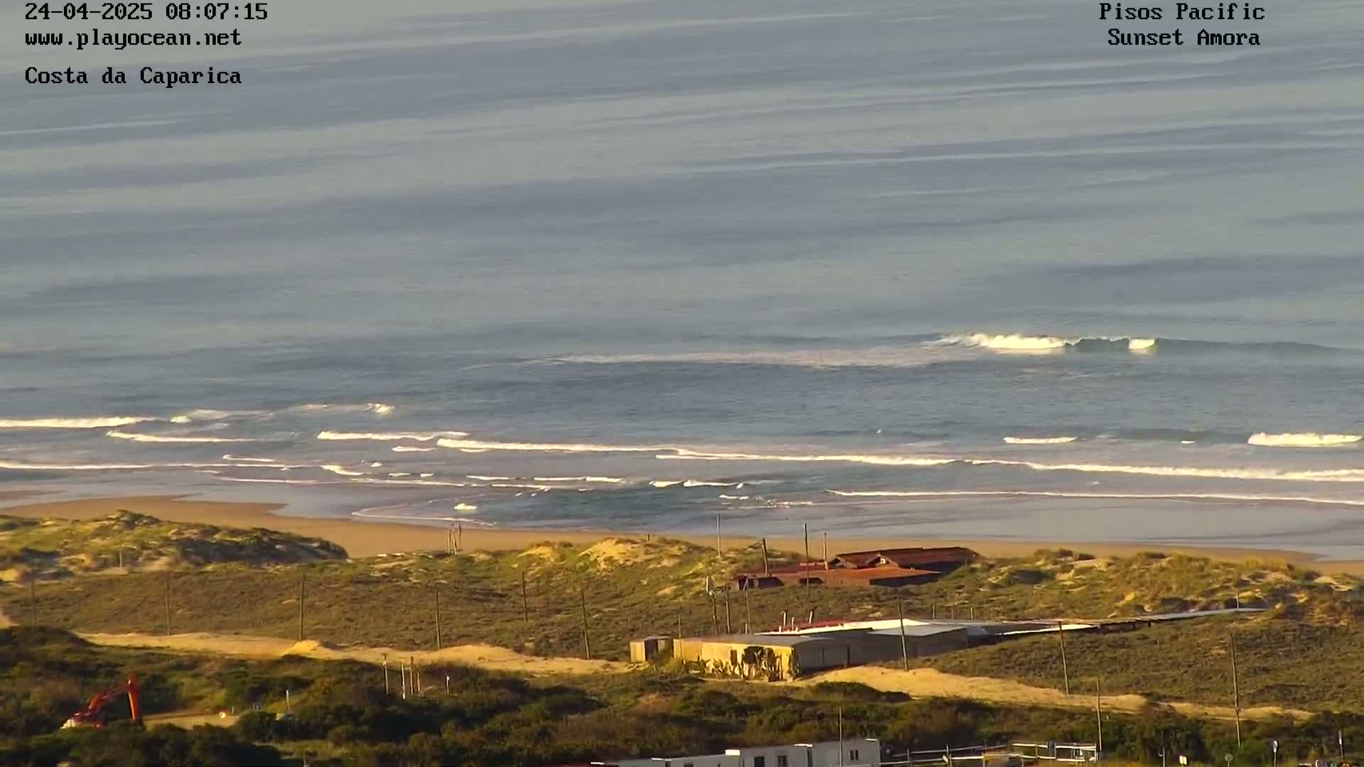 A sandy beach with small waves and dunes is visible, with buildings and scrubland in the foreground under a clear sky.