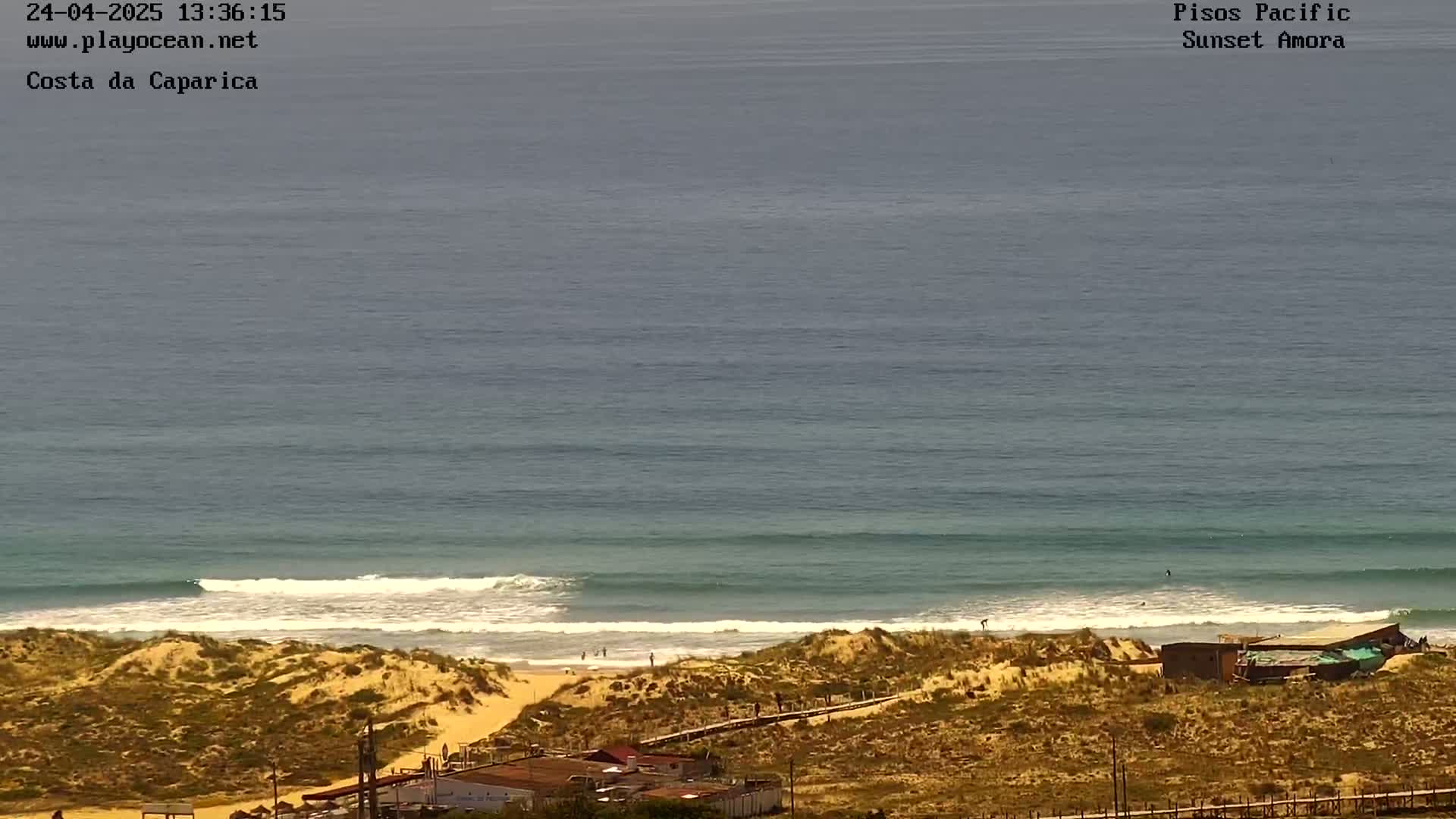 A sandy beach with a few people is visible below a calm ocean with small waves under a clear sky.